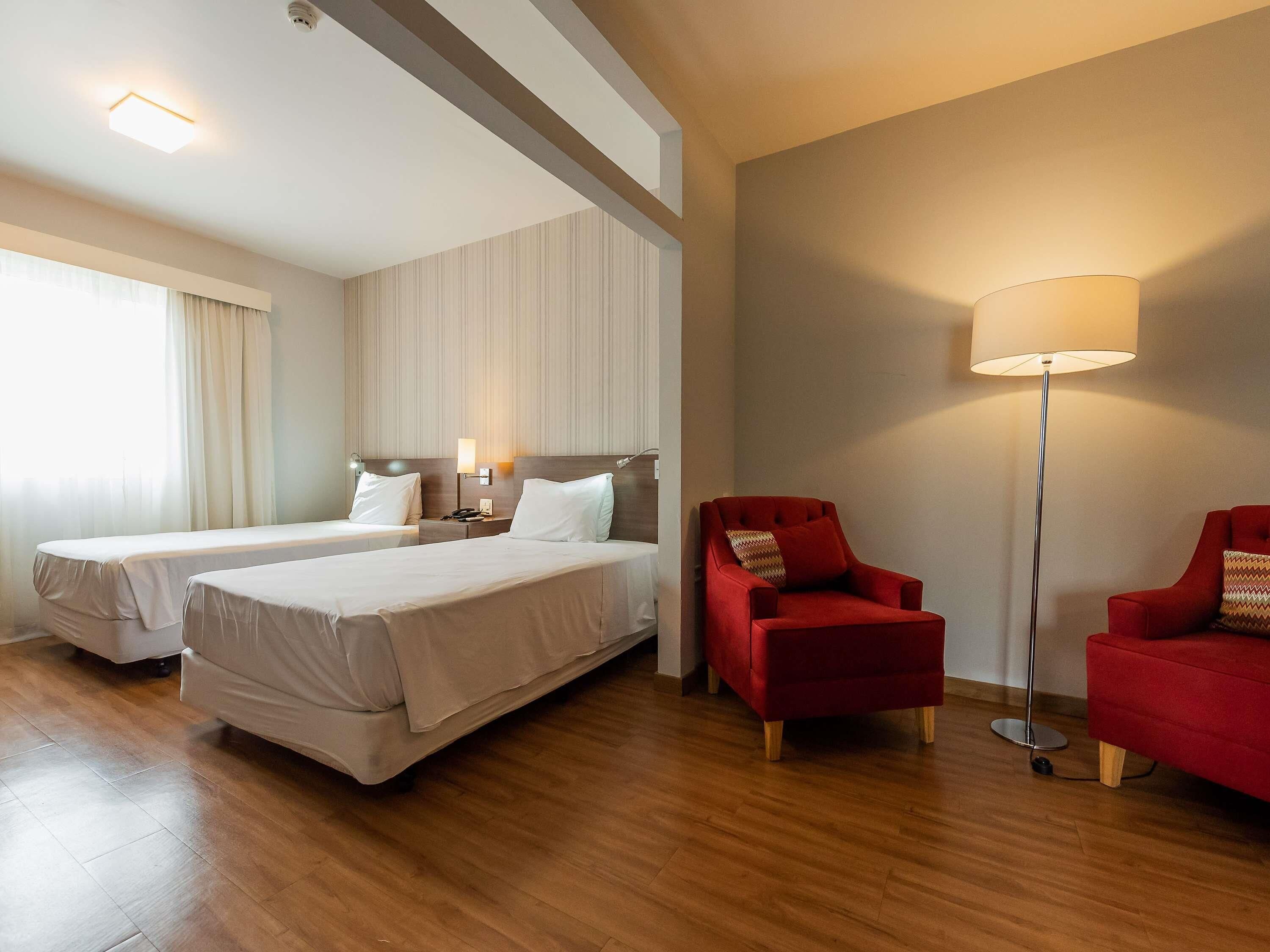 A hotel room with two beds, a red armchair, a floor lamp, and a large window with curtains.