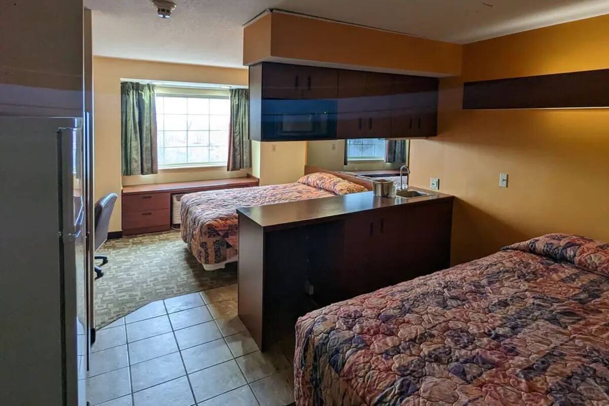 Una habitación de hotel compacta con dos camas individuales, una kitchenette, un pequeño ventanal con cortinas y suelo de baldosas.