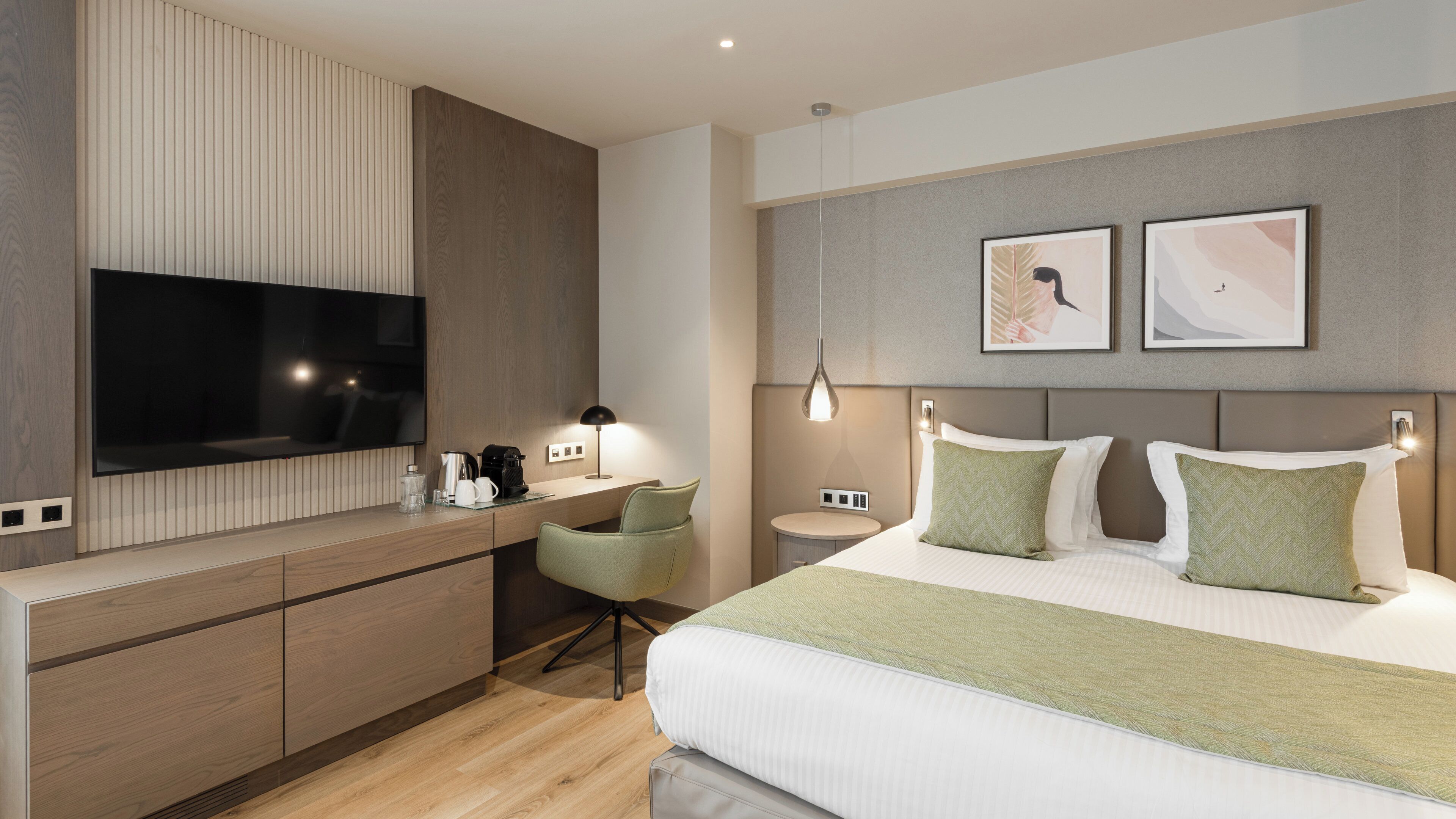 Una habitación de hotel moderna con un televisor de pantalla plana grande, una mesita de noche con lámpara, una mesita redonda, una cama con ropa de cama verde y blanca, y dos obras de arte enmarcadas en la pared.