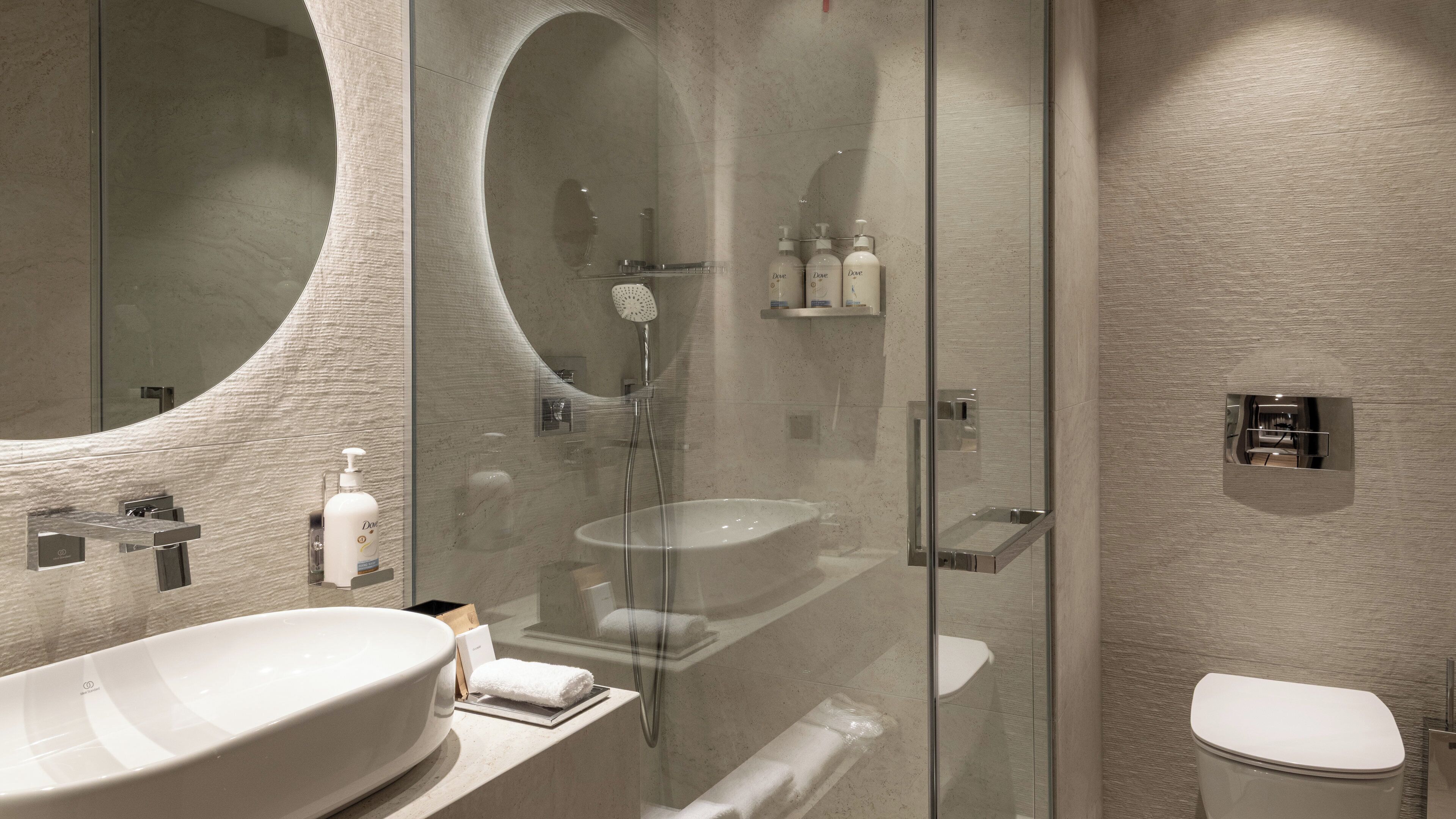 A modern bathroom with a large oval mirror, a glass-enclosed shower, and a white sink.
