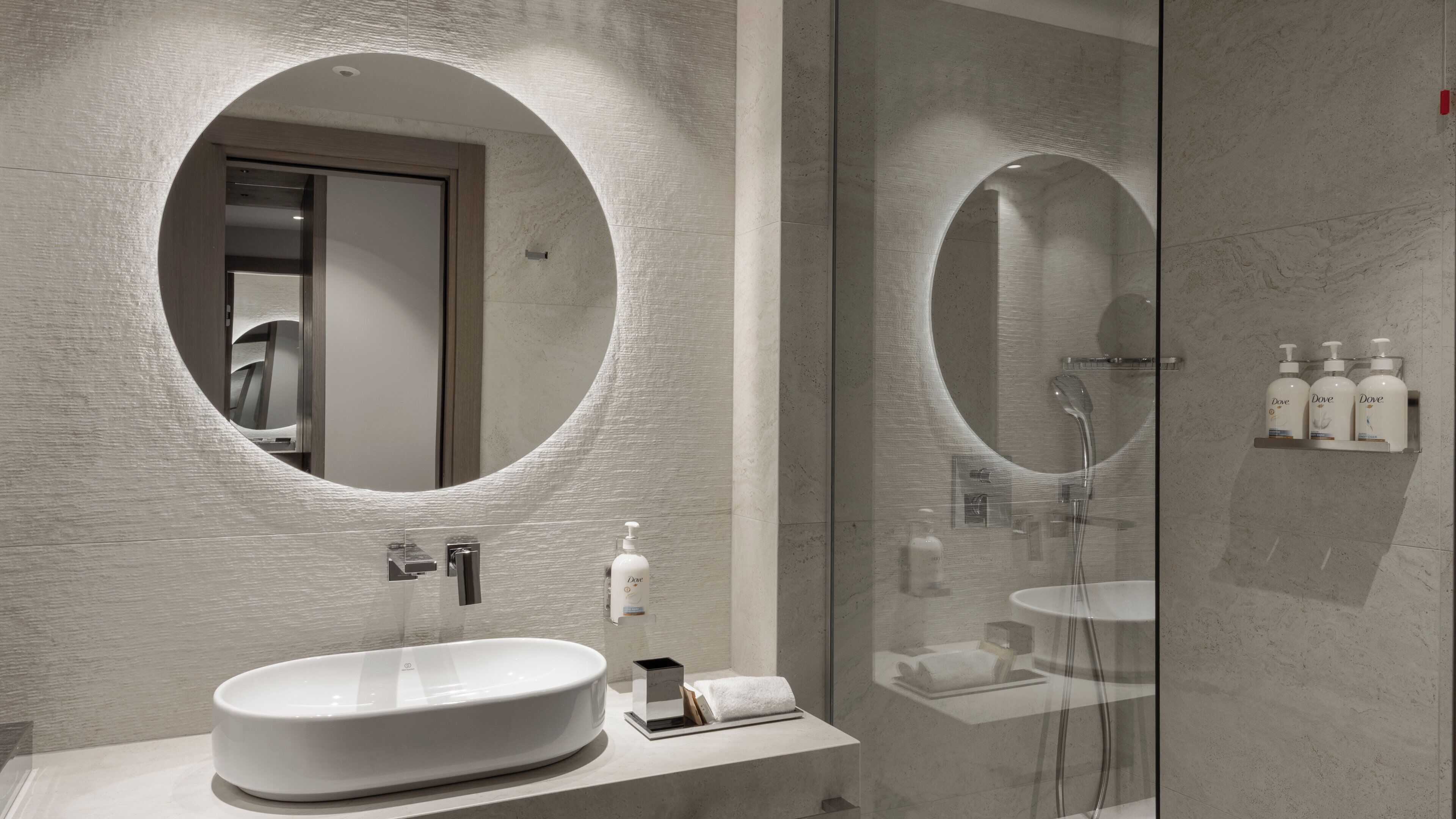Un baño moderno con un lavamanos ovalado grande, un espejo redondo y una zona de ducha con paredes de vidrio.