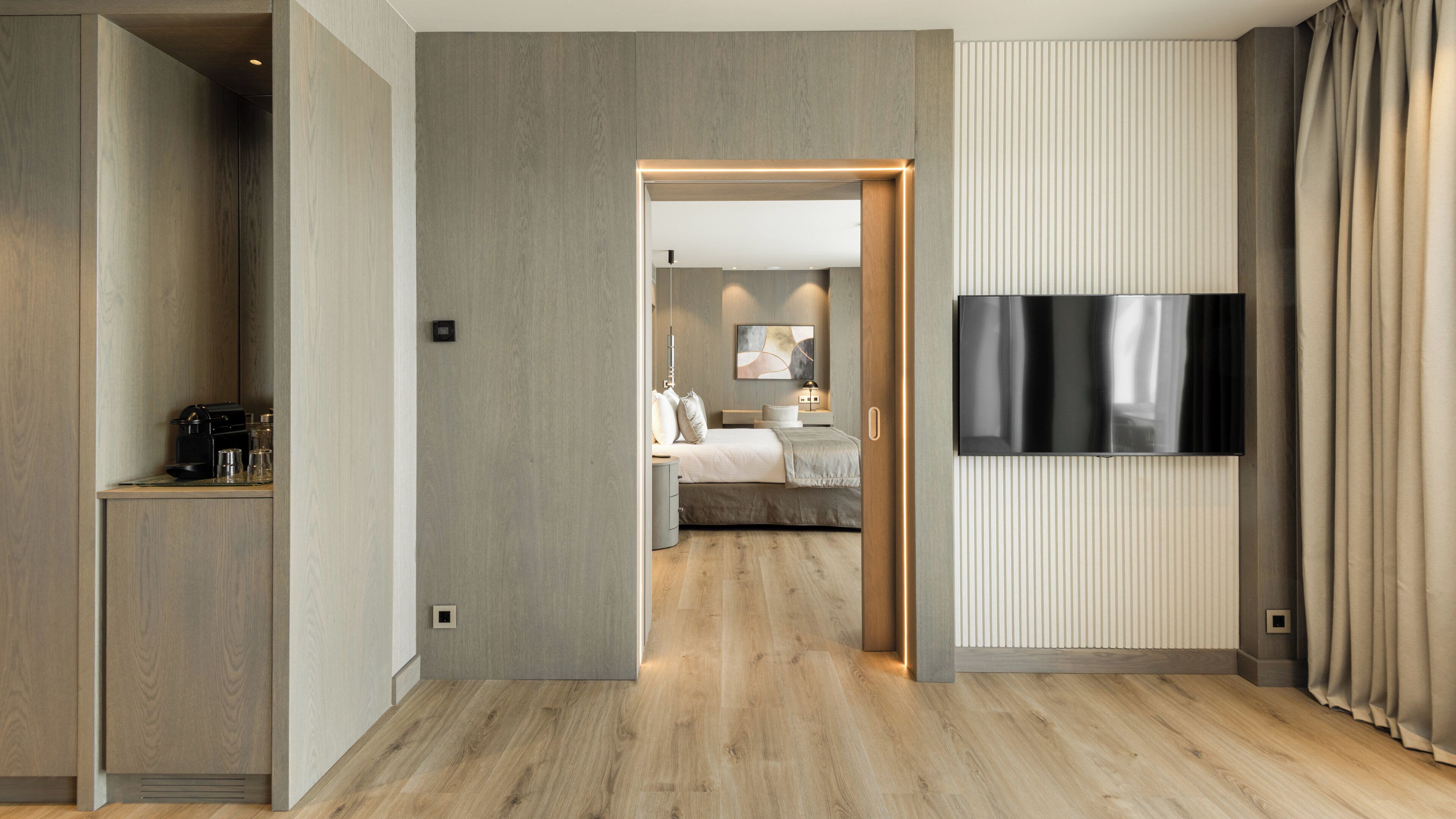 Habitación de hotel con piso de madera, una cama con cabecera, una mesita de noche, un televisor de pantalla plana montado en la pared y vista a la habitación contigua.