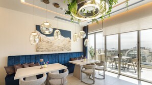 A modern dining area with a blue upholstered bench, a white table, and four chairs. There are pendant lights and a large window offering a city view. - Holiday Inn Thessaloniki by IHG (Thessaloniki)