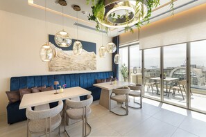 A modern dining area with a blue upholstered bench, a white table, and four chairs. There are pendant lights and a large window offering a city view. - Holiday Inn Thessaloniki by IHG (Thessaloniki)