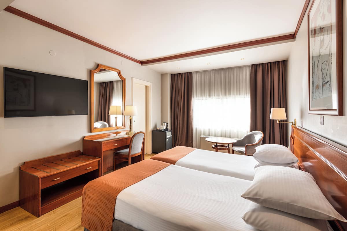 A hotel room with two beds, a desk, a chair, a TV, a mirror, and a window with curtains.