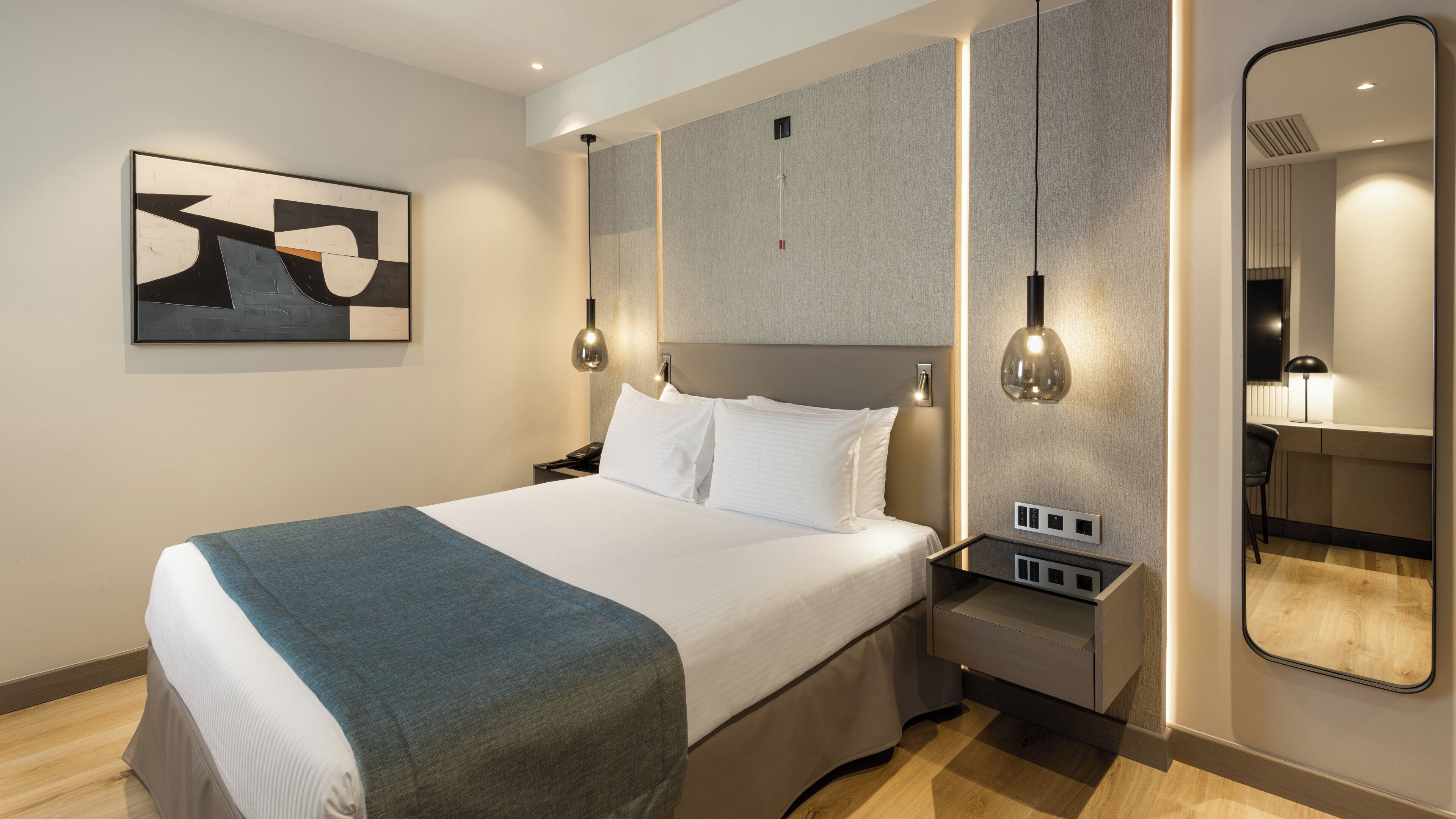 A modern hotel room with a large bed, bedside tables, a wall-mounted artwork, and a mirror.