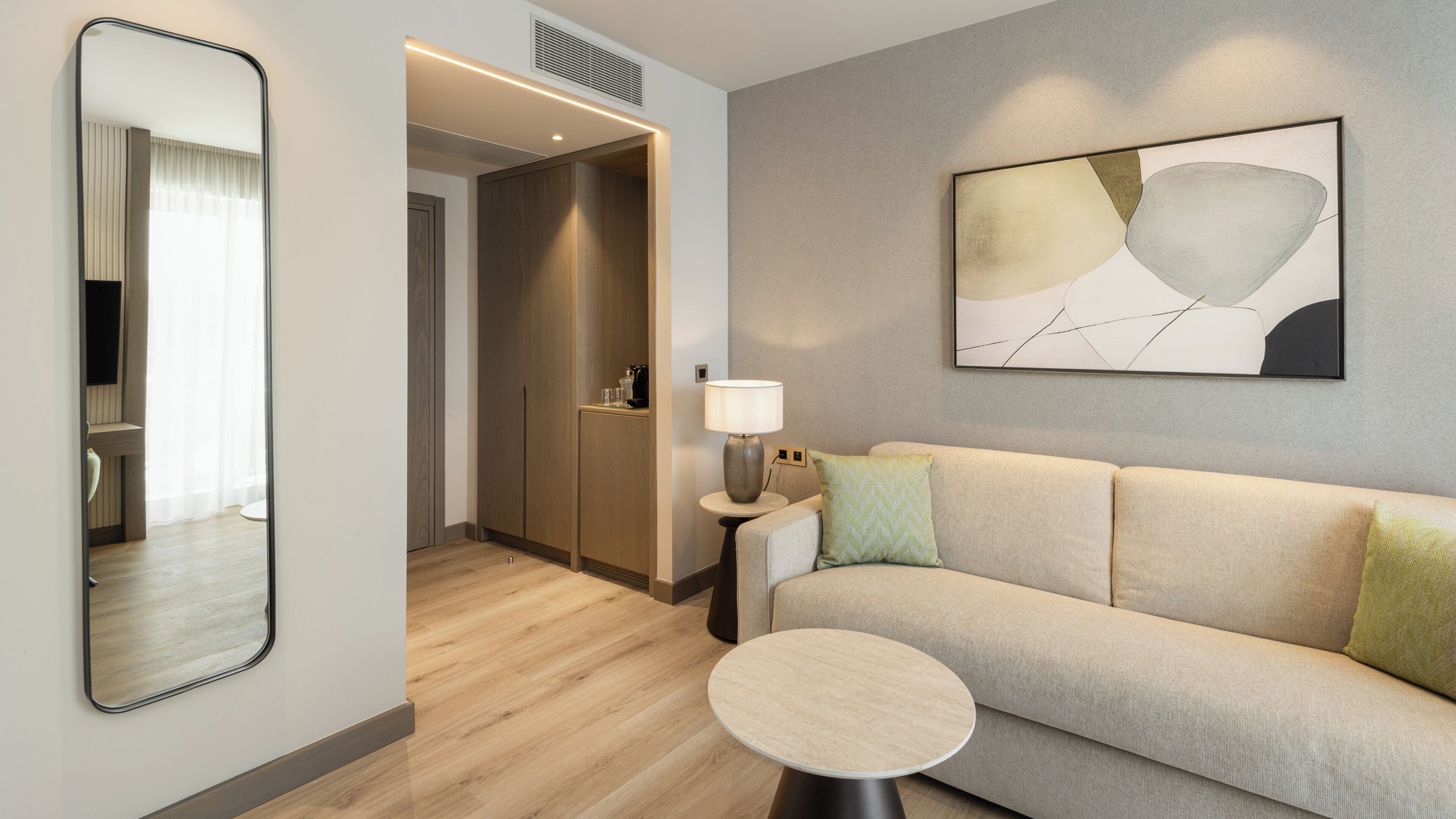 A modern hotel room with a sofa, a round table, a lamp, a painting, and a mirror.