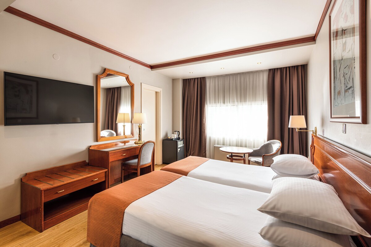 A hotel room with two beds, a desk, a chair, a TV, a mirror, and a window with curtains.