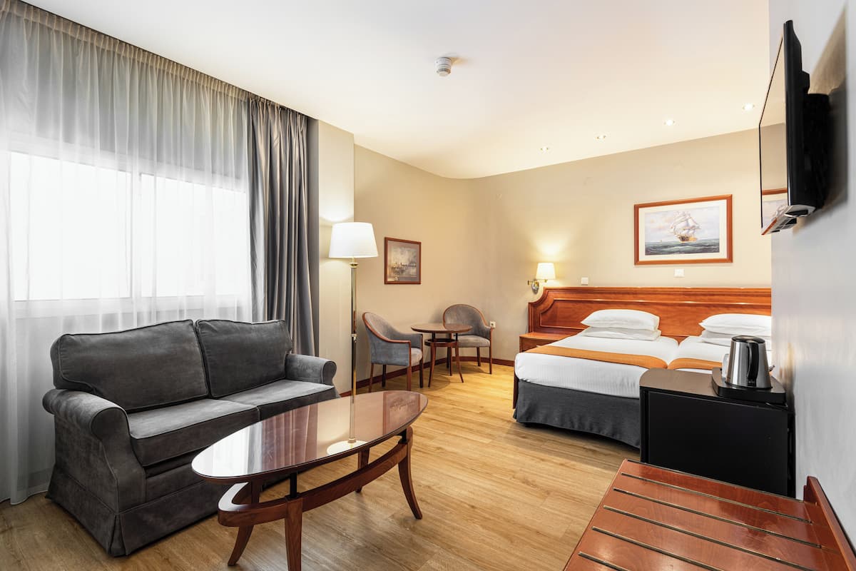 A hotel room with a sofa, a small table, a bed with two pillows, a nightstand, a lamp, and a painting on the wall.