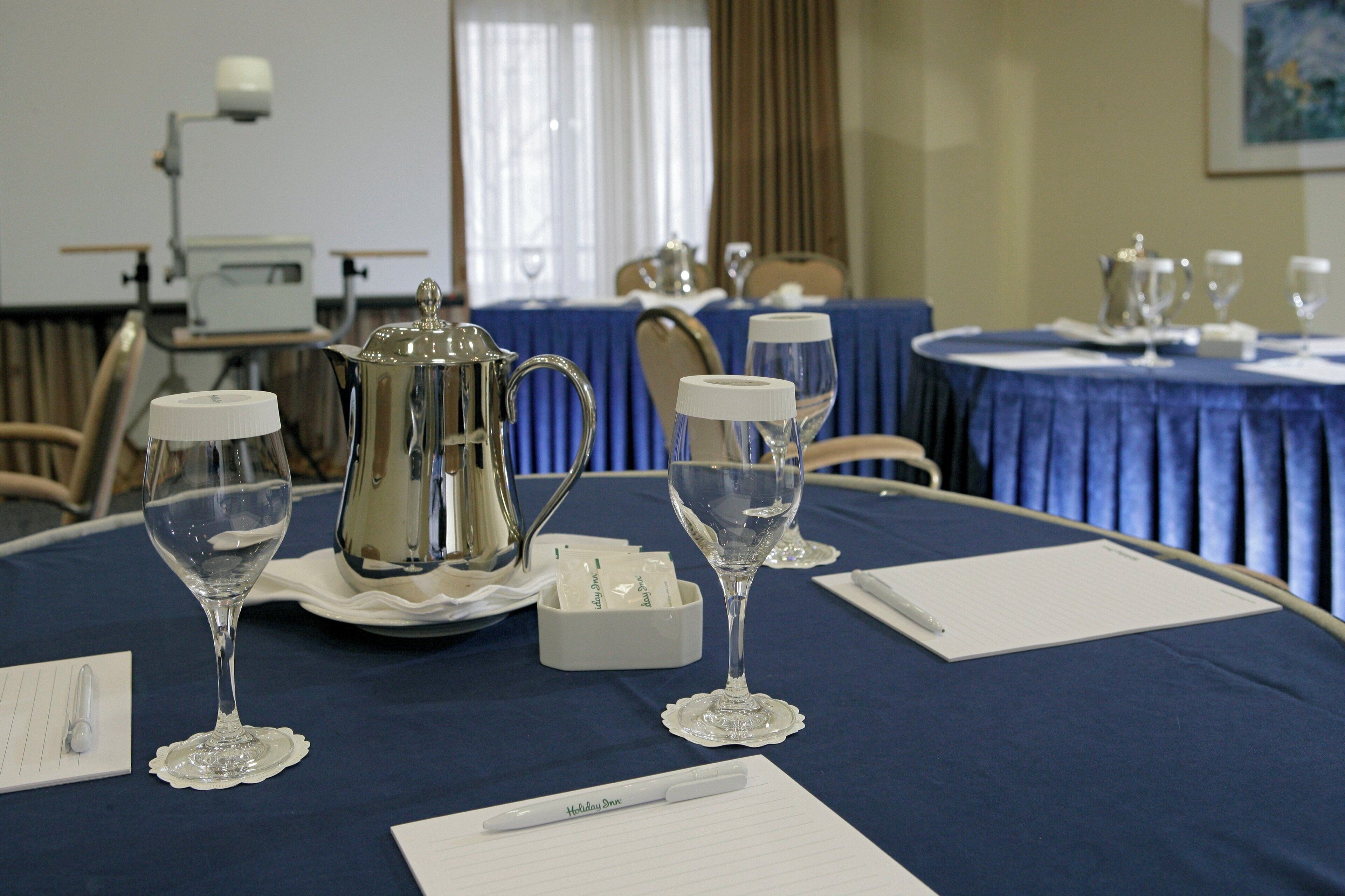 Un salón de conferencias con mesas dispuestas para una reunión, equipado con tarjetas de identificación, blocs de notas, bolígrafos y copas.