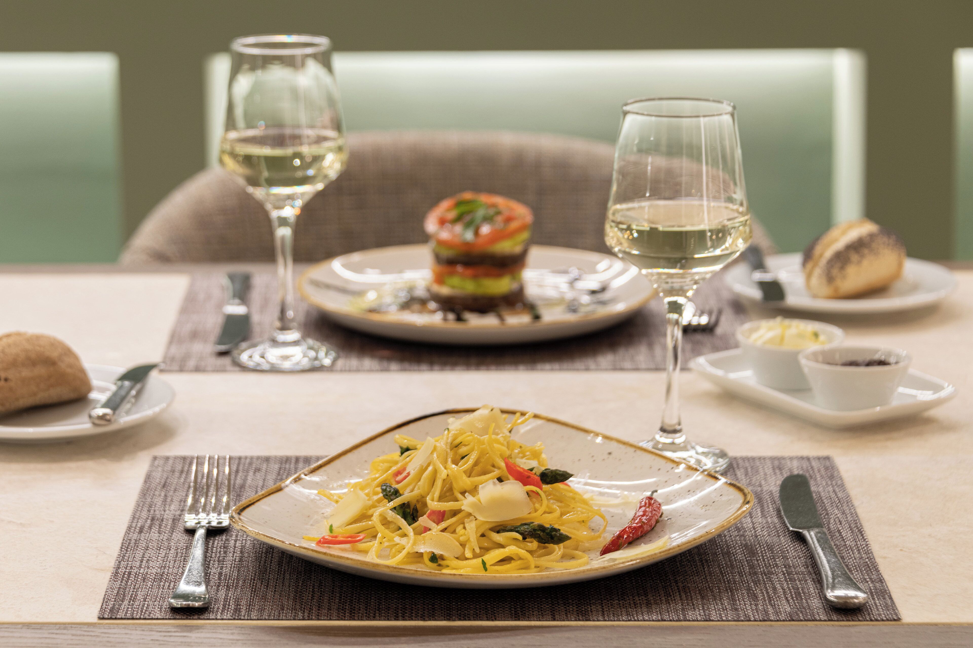 A dining table with plates of pasta, a burger, and a glass of white wine.