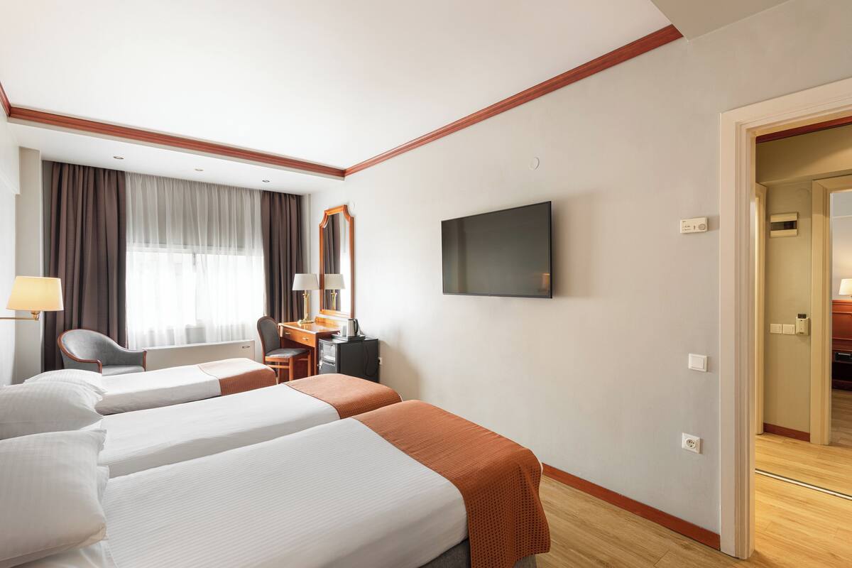 A hotel room with two beds, a desk, a chair, a TV, and a large window with curtains.