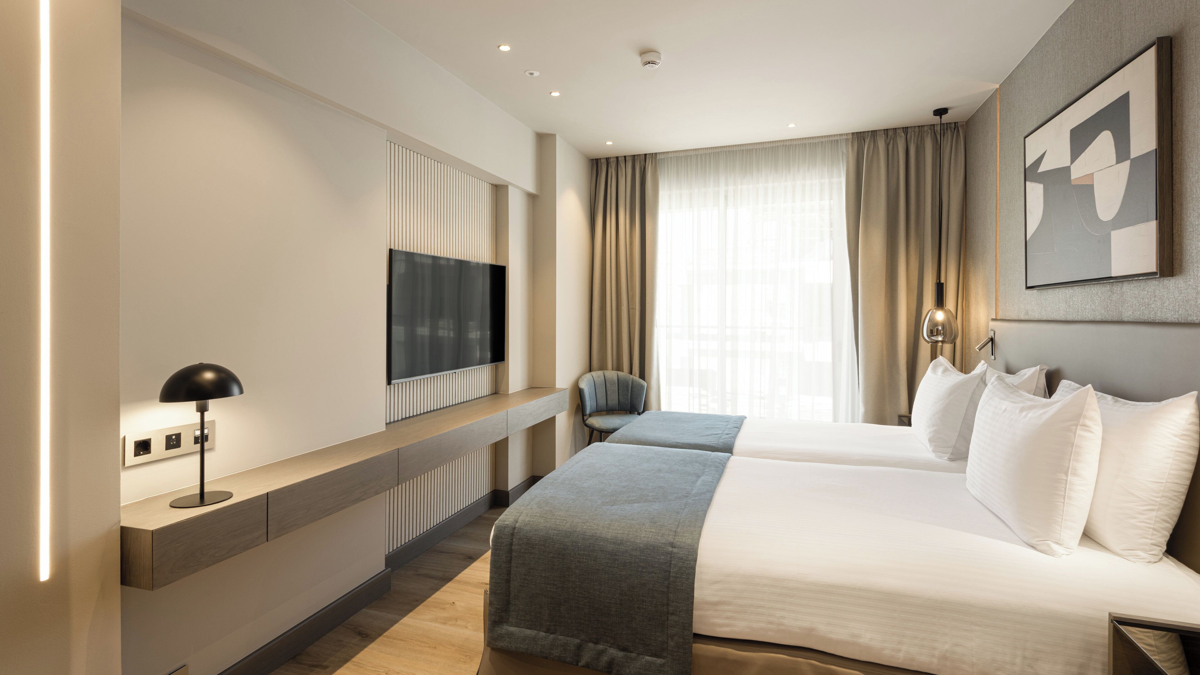 A hotel room with a large bed, a TV mounted on the wall, a desk with a lamp, and a window with curtains.