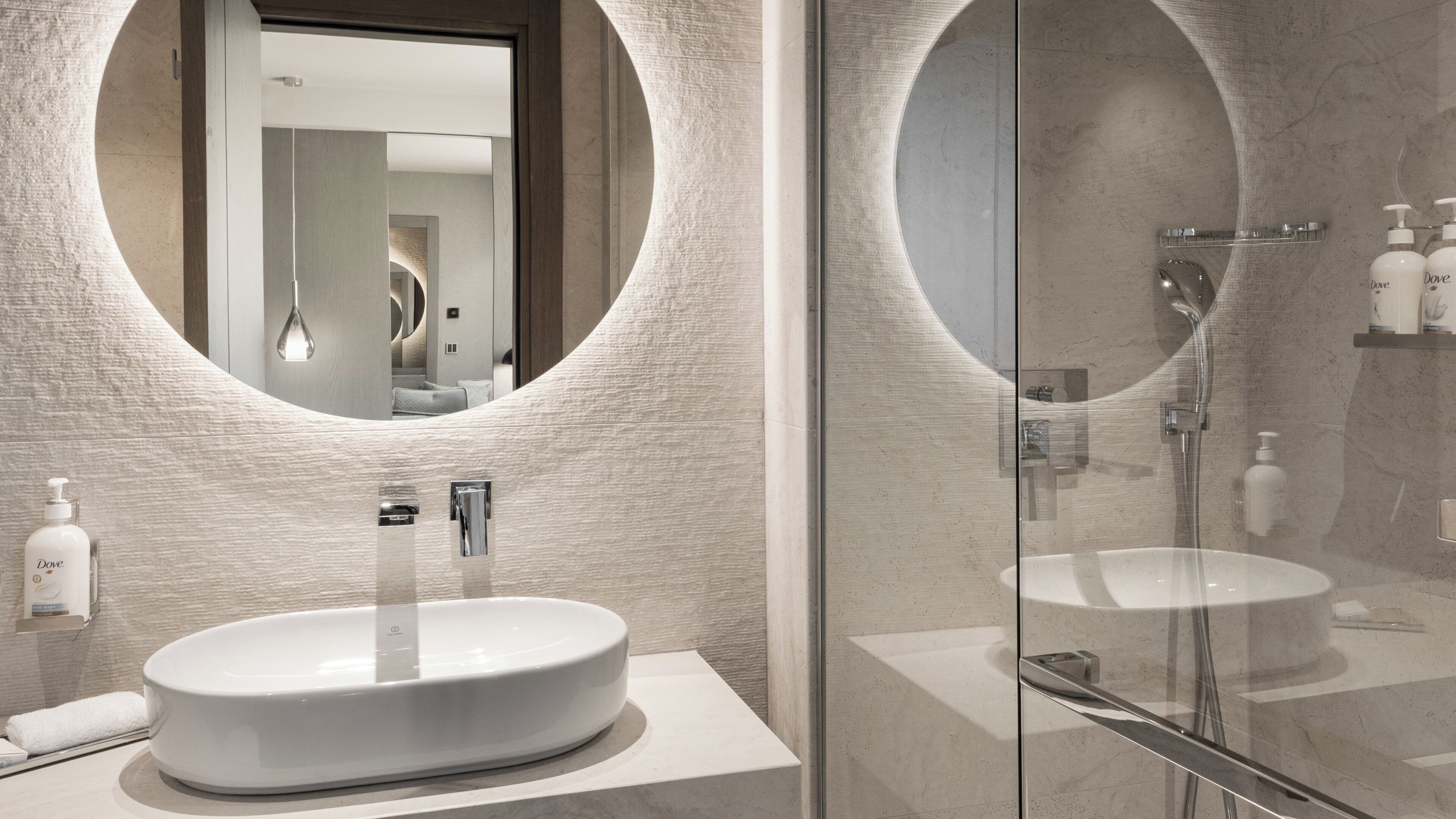 Een moderne badkamer met een grote spiegel in ovaalvorm, een ronde wasbak en een douchecabine met glazen wanden.