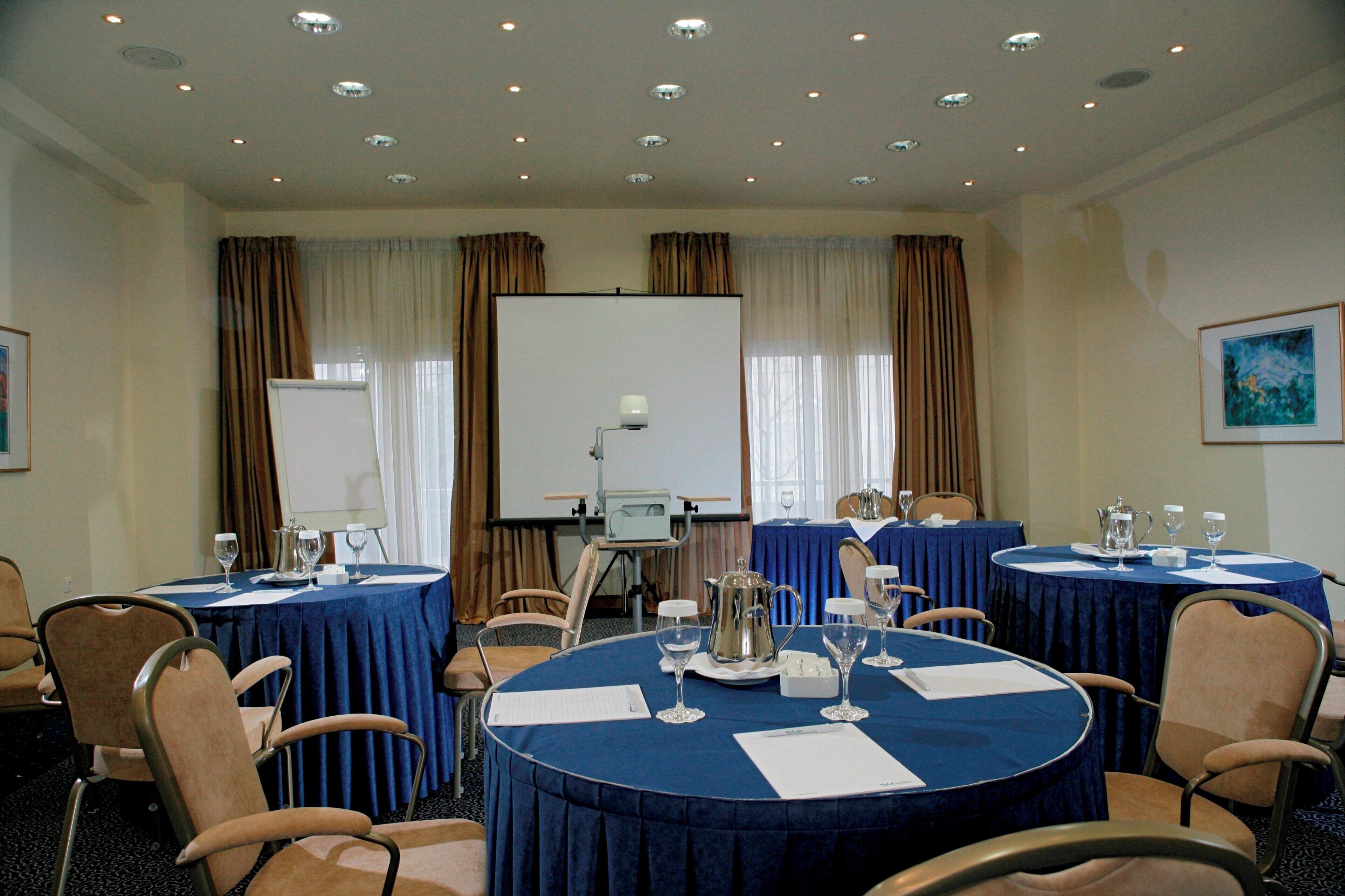 Una sala de conferencias con mesas redondas, sillas, una pantalla de proyector y una pizarra.