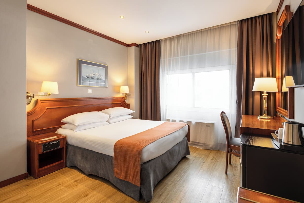 A hotel room with a large bed, bedside lamps, a desk with a chair, and a window with curtains.