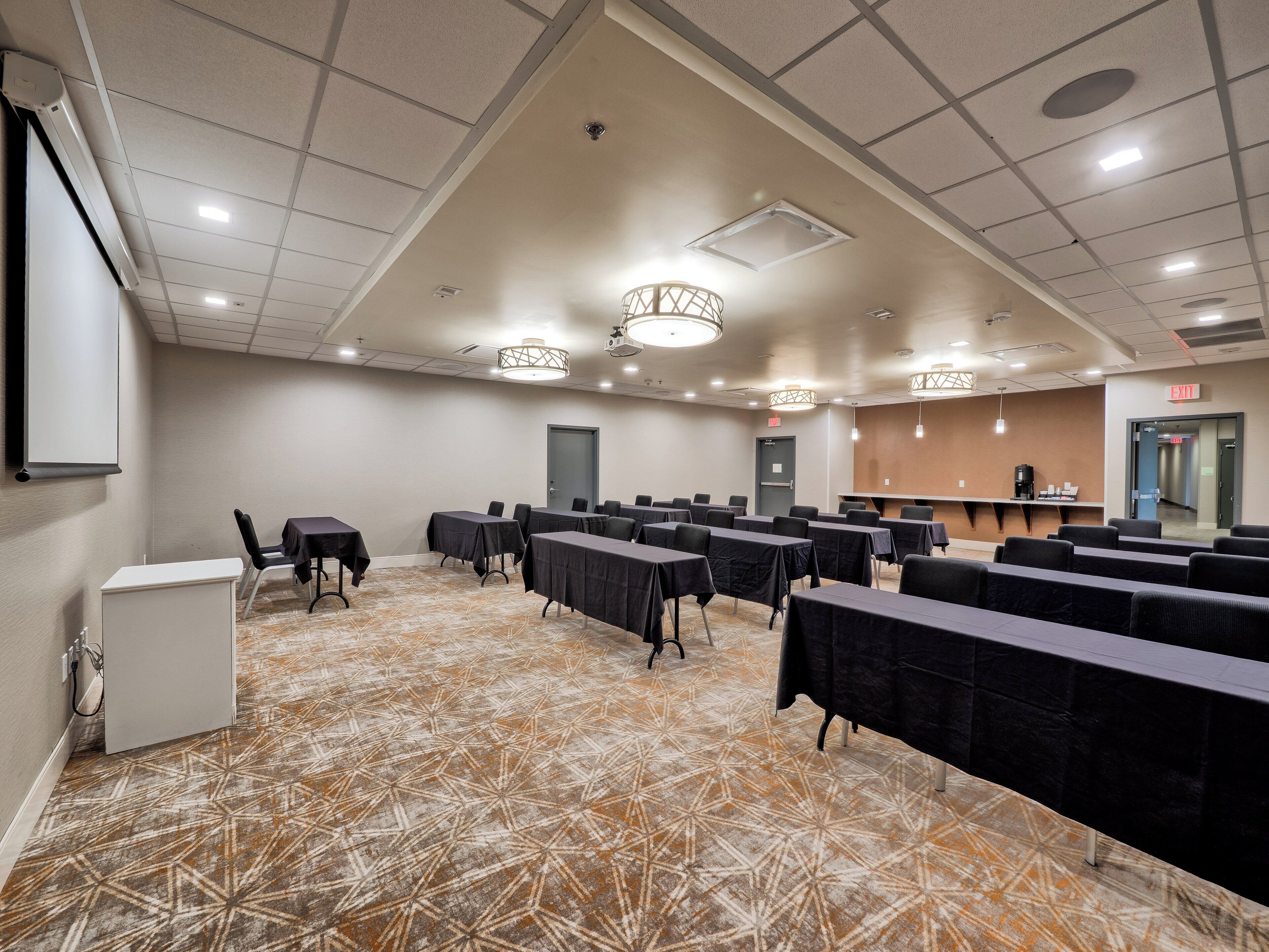 Une salle de conférence équipée de tables, de chaises, d’un écran de projection, d’un coin café et d’une moquette à motifs.