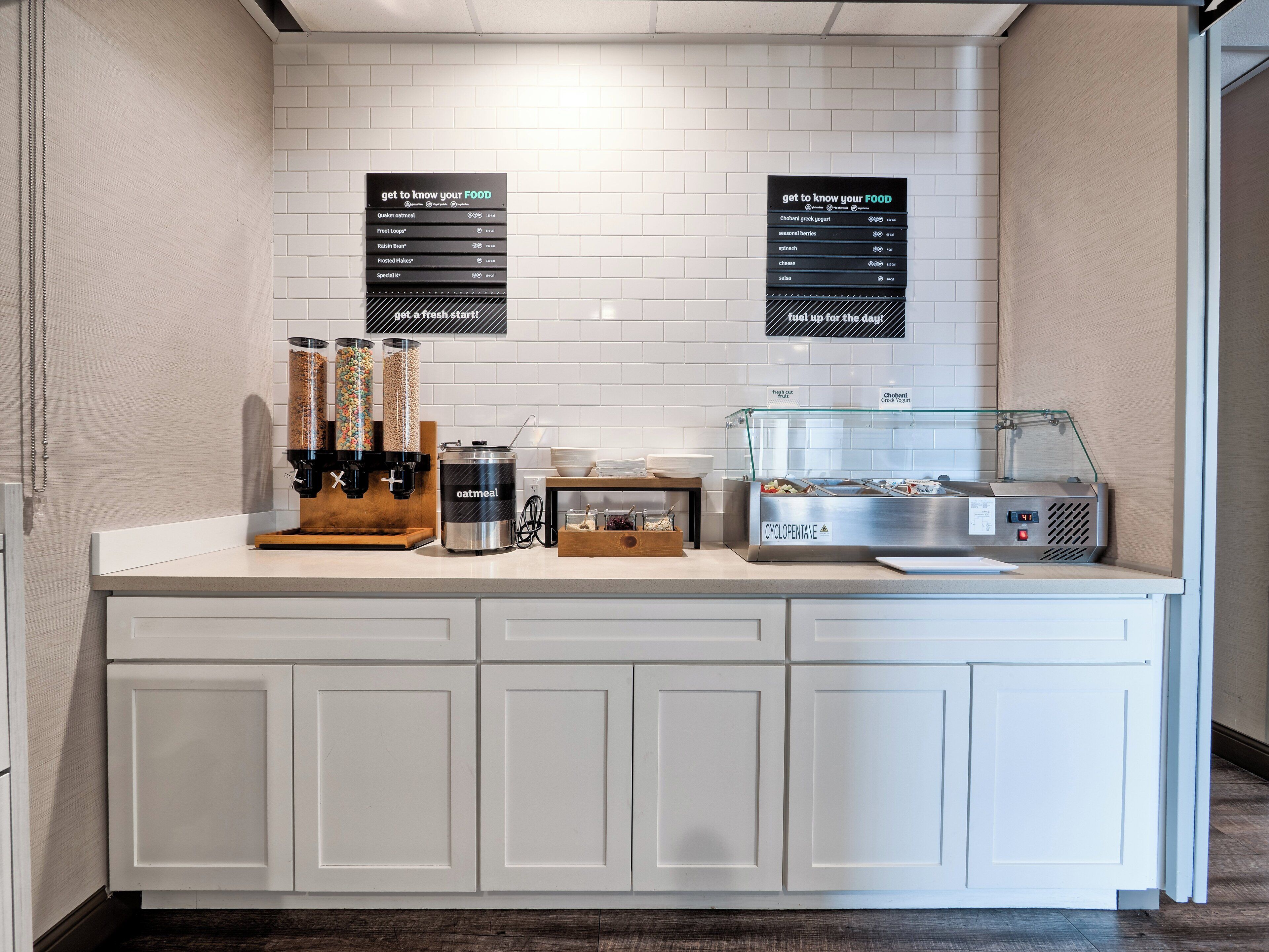 Ein Frühstücksbuffet mit Kaffeemaschine, Obstspender und einem Kühlschrank für Joghurt.