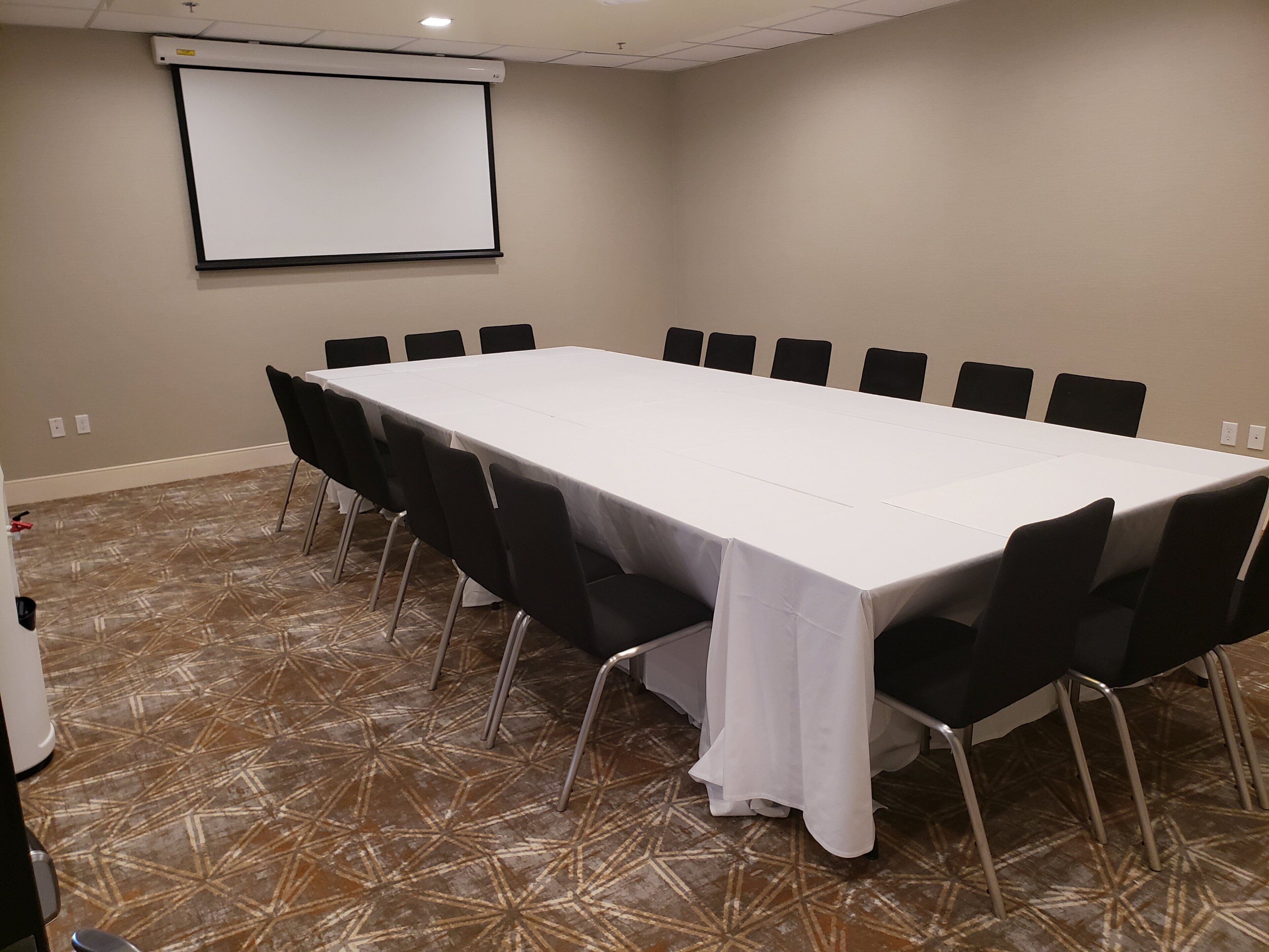 Une salle de conférence avec une longue table, des chaises noires, un écran de projection et une moquette à motifs.
