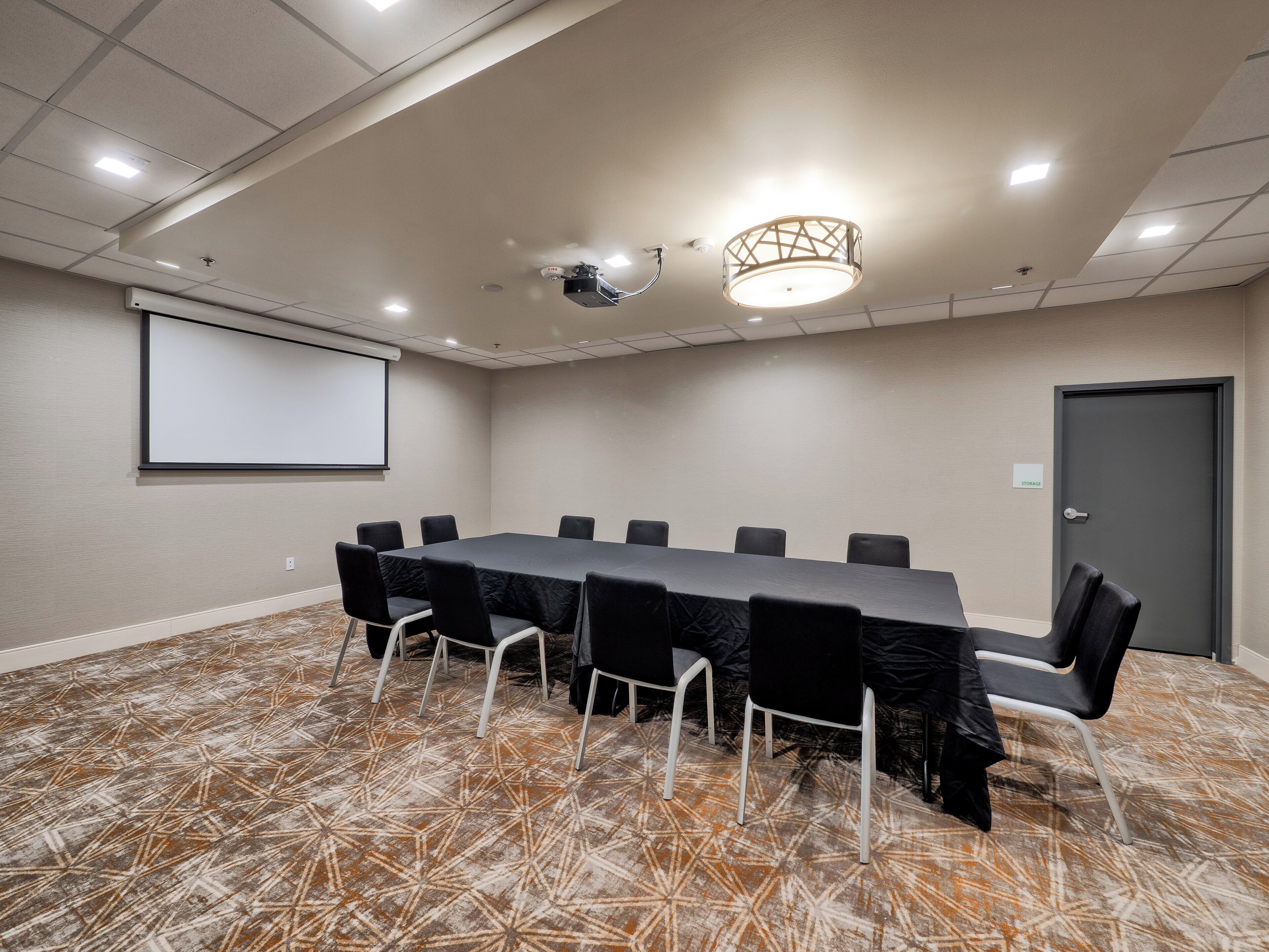 A conference room with a long table, black chairs, a projector screen, and a patterned carpet.