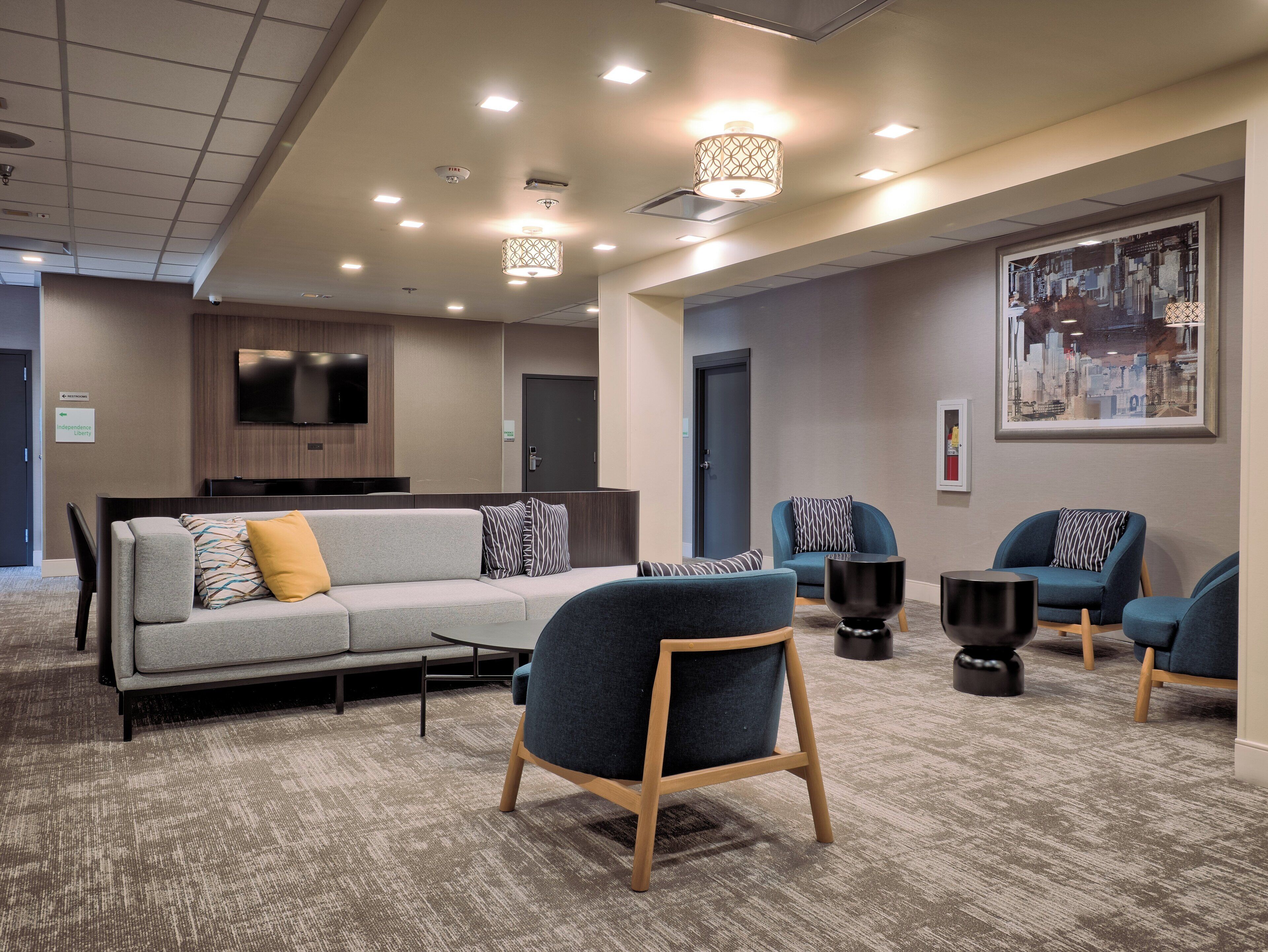 A modern hotel lobby with a grey sofa, blue armchairs, a flat-screen TV, and a cityscape painting.