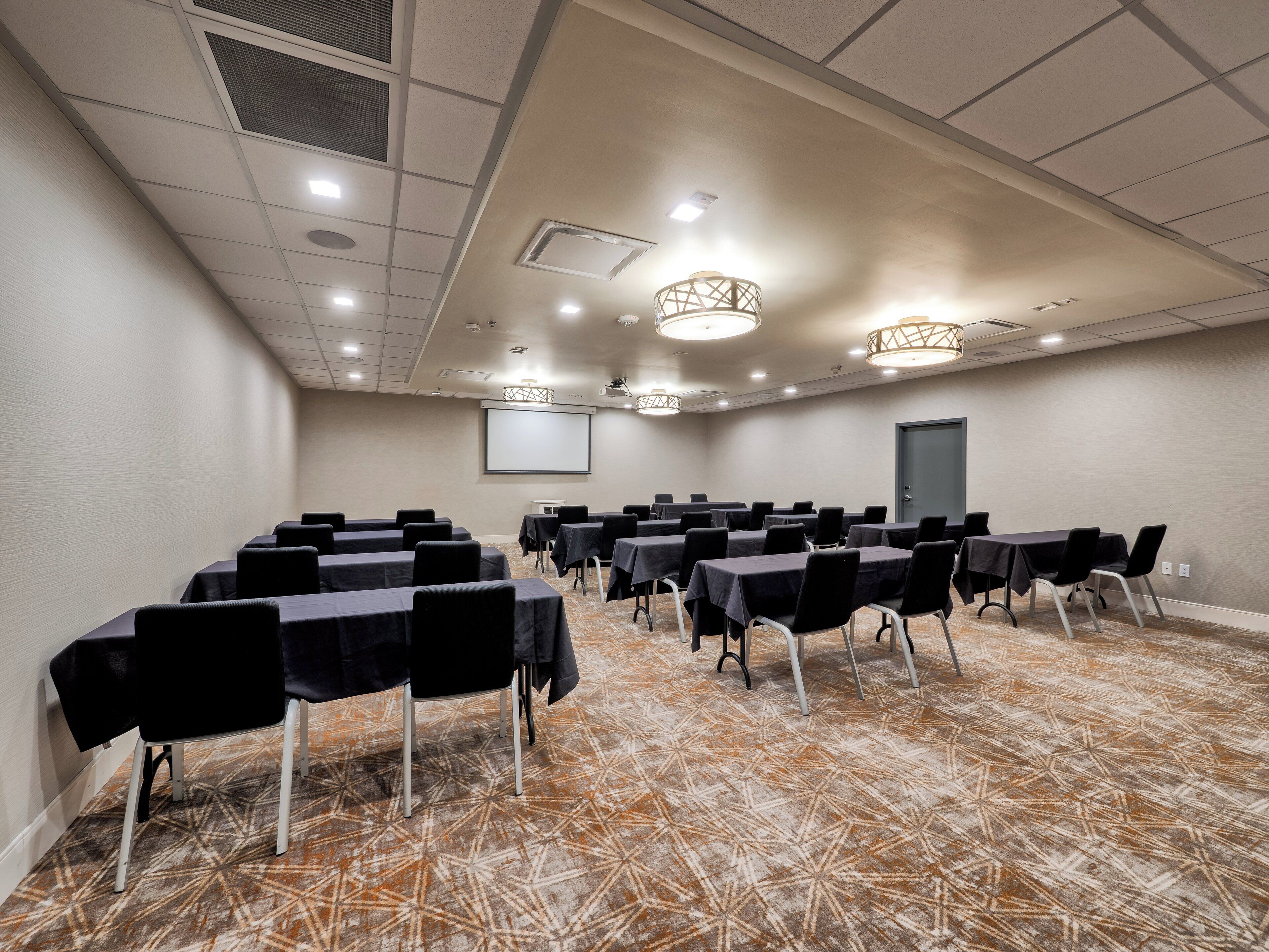 A conference room with rows of tables and chairs, a projector screen, and a patterned carpet.
