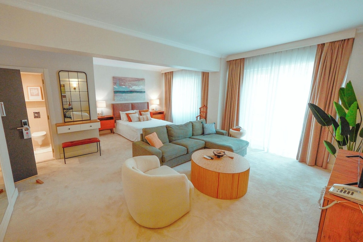 A hotel room with a bed, a sofa, a round coffee table, and a large window with curtains.