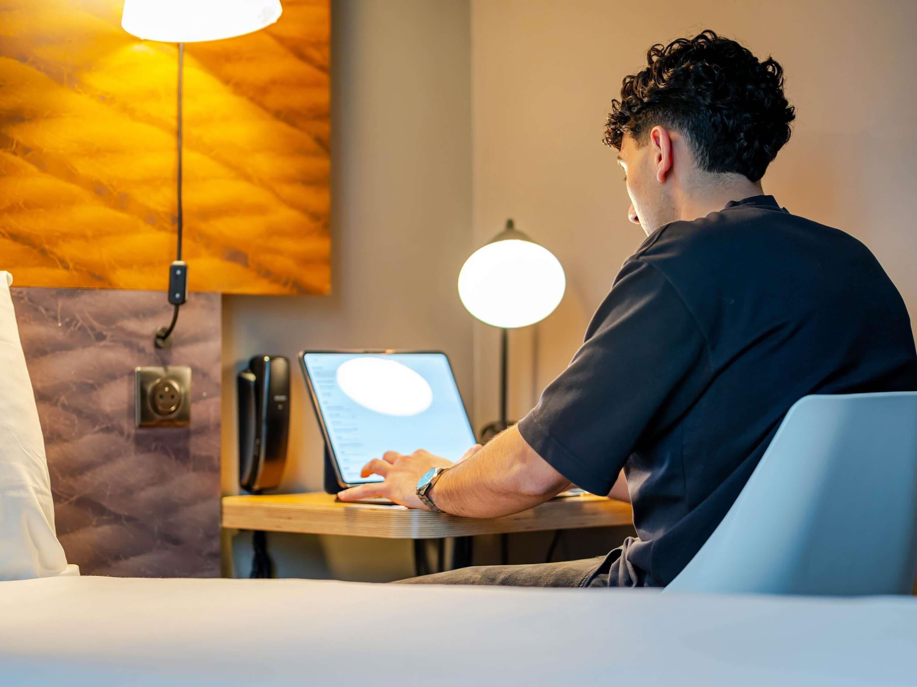 A person using a laptop at a wooden desk in a room with a marble wall and pendant lighting.