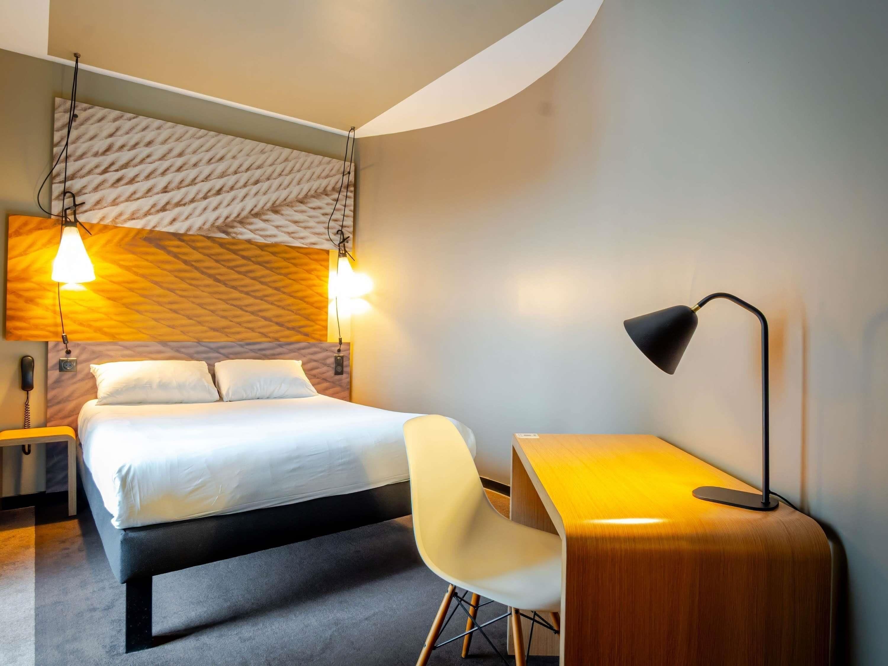 A modern hotel room with a bed, bedside lamp, desk, chair, and a patterned headboard.