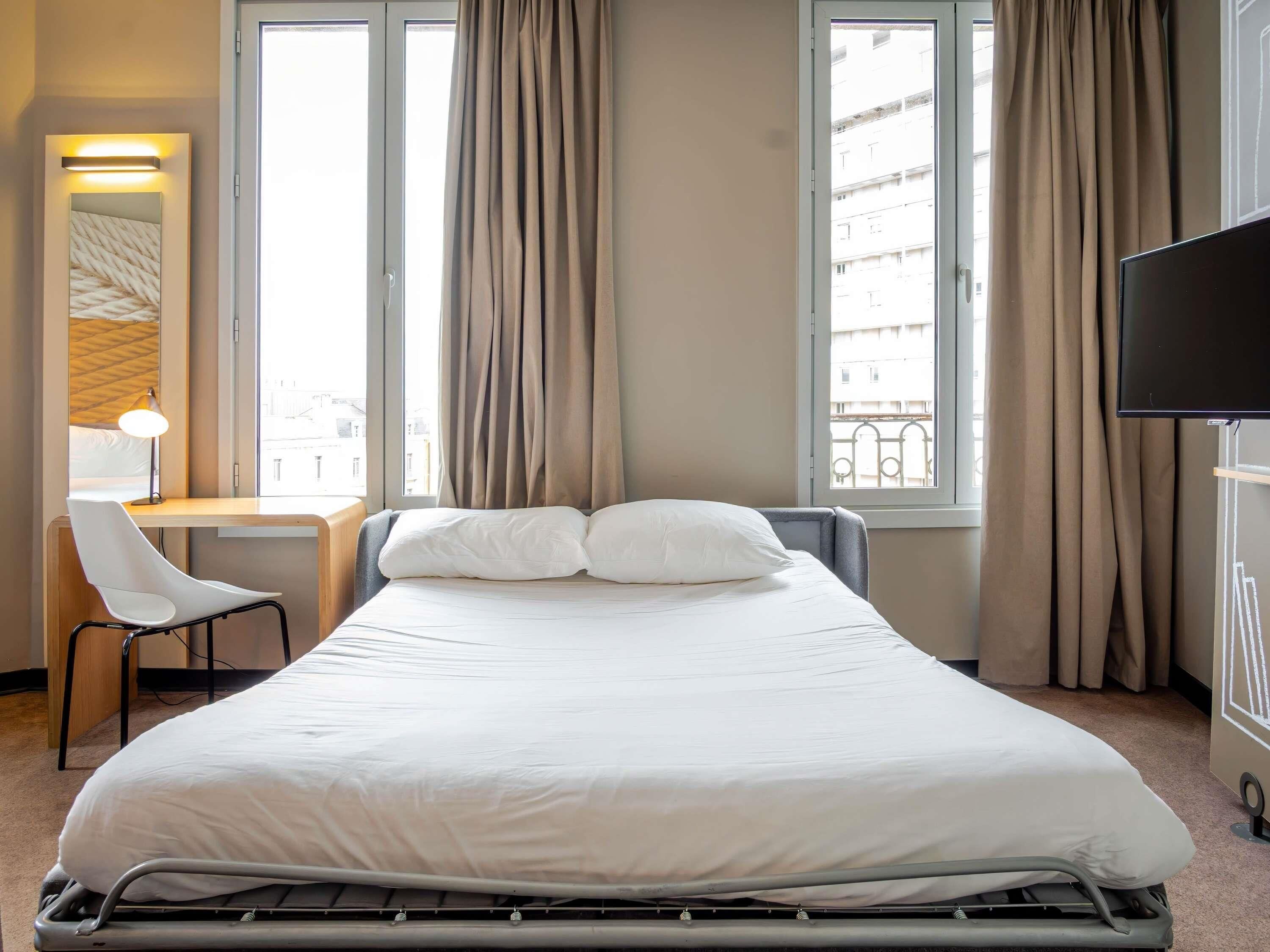 A hotel room with a bed, a desk, a chair, a TV, and large windows with curtains.