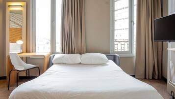 A hotel room with a bed, a desk, a chair, a TV, and large windows with curtains.