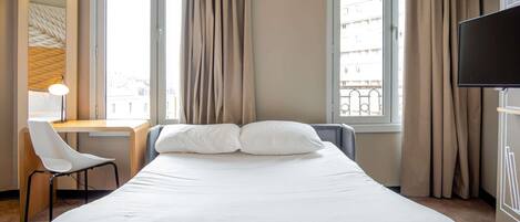 A hotel room with a bed, a desk, a chair, a TV, and large windows with curtains.