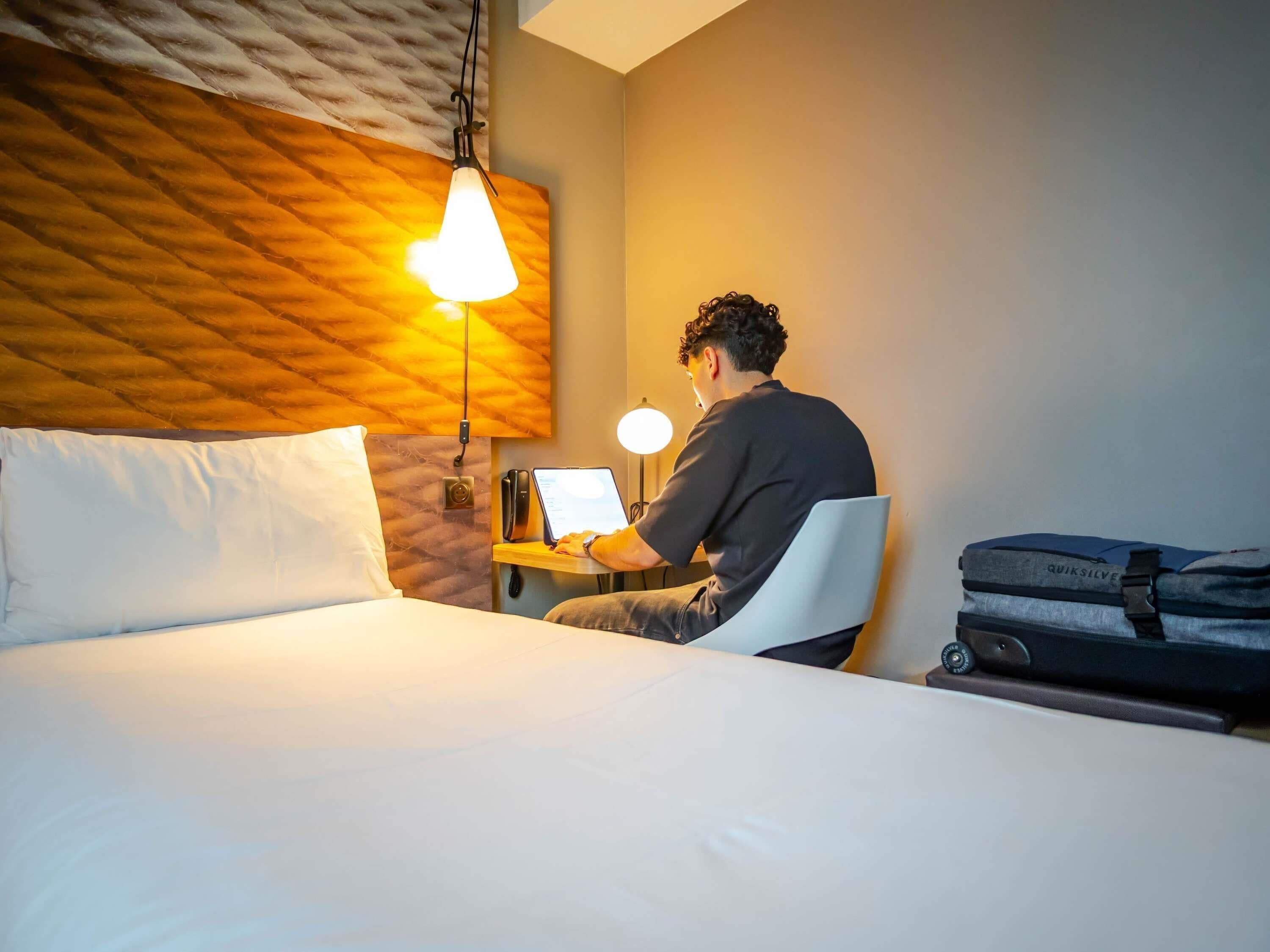 A hotel room with a bed, a desk with a laptop, a desk lamp, and a suitcase.