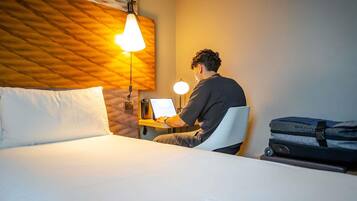 A hotel room with a bed, a desk with a laptop, a desk lamp, and a suitcase.