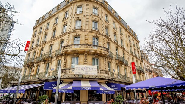 Un edificio storico con un ingresso ad angolo, una pizzeria ristorante chiamata 'Terminus' con tavoli all'aperto e un cartello del 'Tis Hotel'.