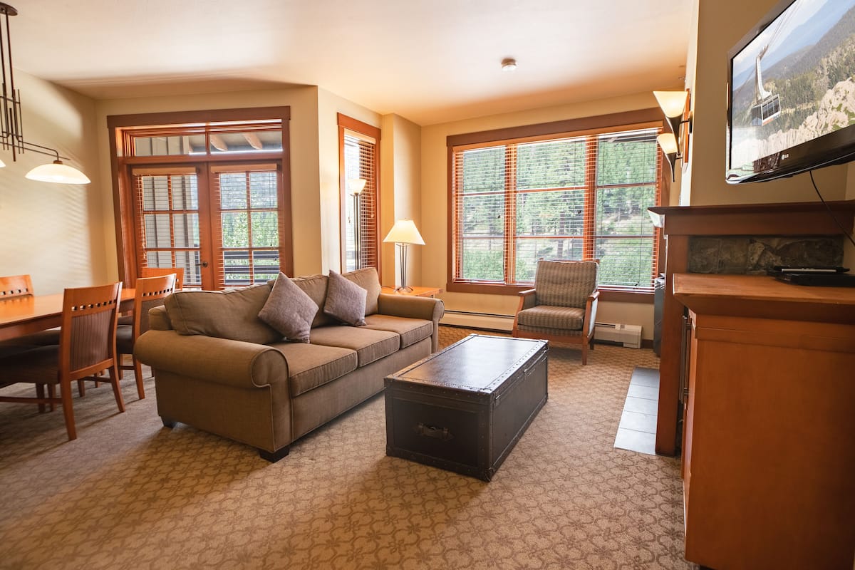 A living room with a sofa, coffee table, and TV. There are windows with blinds, a lamp, and a fireplace.