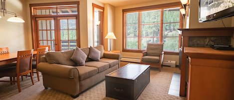 A living room with a sofa, coffee table, and TV. There are windows with blinds, a lamp, and a fireplace.
