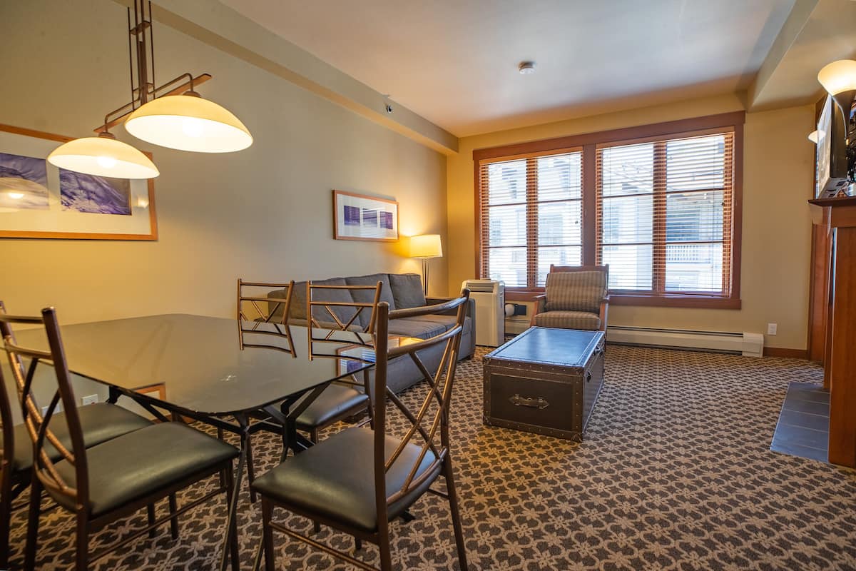 A hotel room with a dining table, chairs, a sofa, a small cabinet, and a window with blinds.