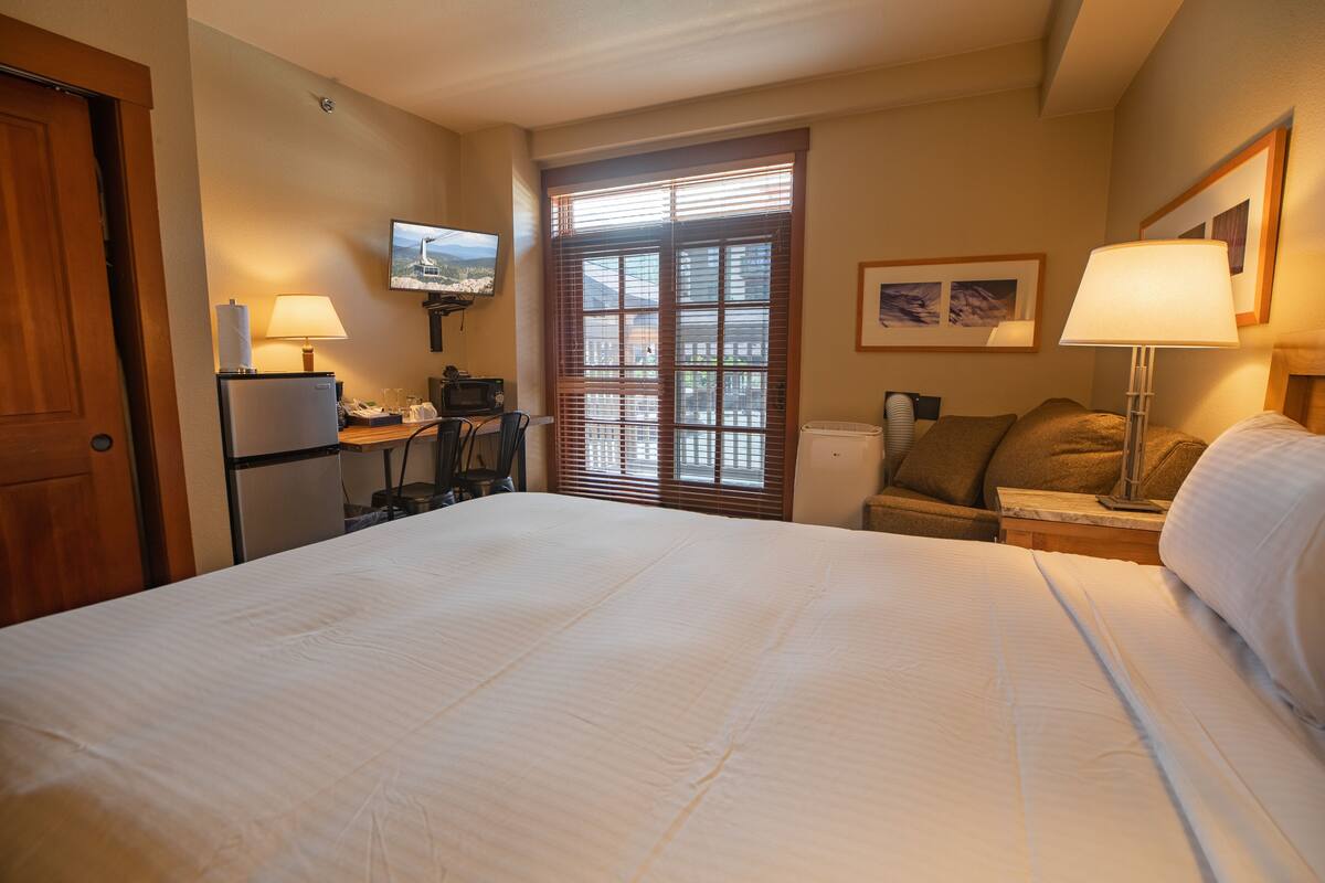 A hotel room with a large bed, a desk with a computer, a refrigerator, a microwave, a chair, a window with blinds, and a television on the wall.