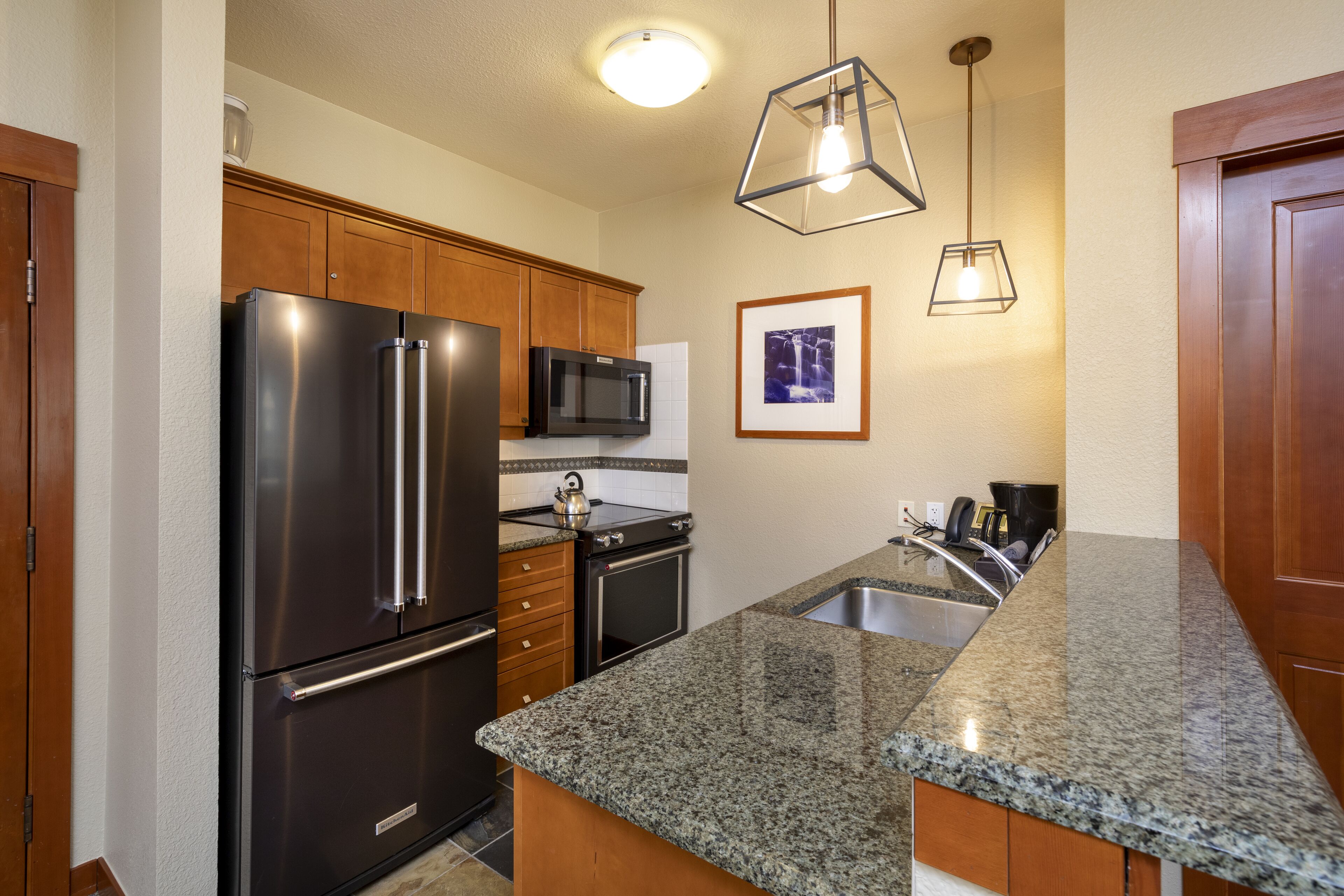A kitchen with granite countertops, stainless steel appliances, wooden cabinets, a microwave, a kettle, and pendant lighting.