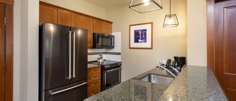 A kitchen with granite countertops, stainless steel appliances, wooden cabinets, a microwave, a kettle, and pendant lighting.