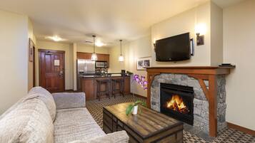 A living room with a fireplace, a sofa, a coffee table, and a kitchen in the background.
