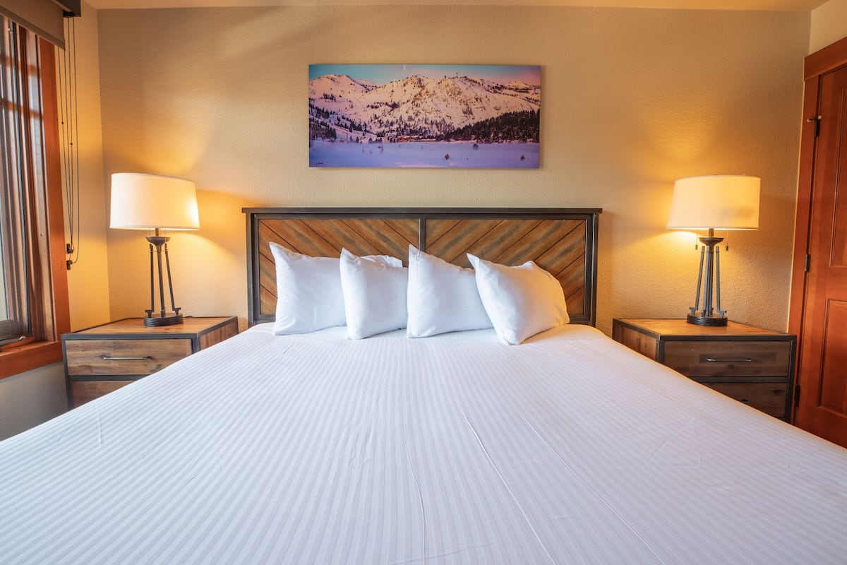 A hotel room with a large bed, two bedside tables with lamps, a wooden headboard, and a mountain landscape painting on the wall.