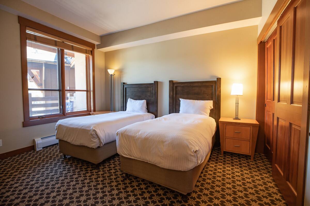 A hotel room with two beds, a nightstand, a lamp, and a window with blinds.