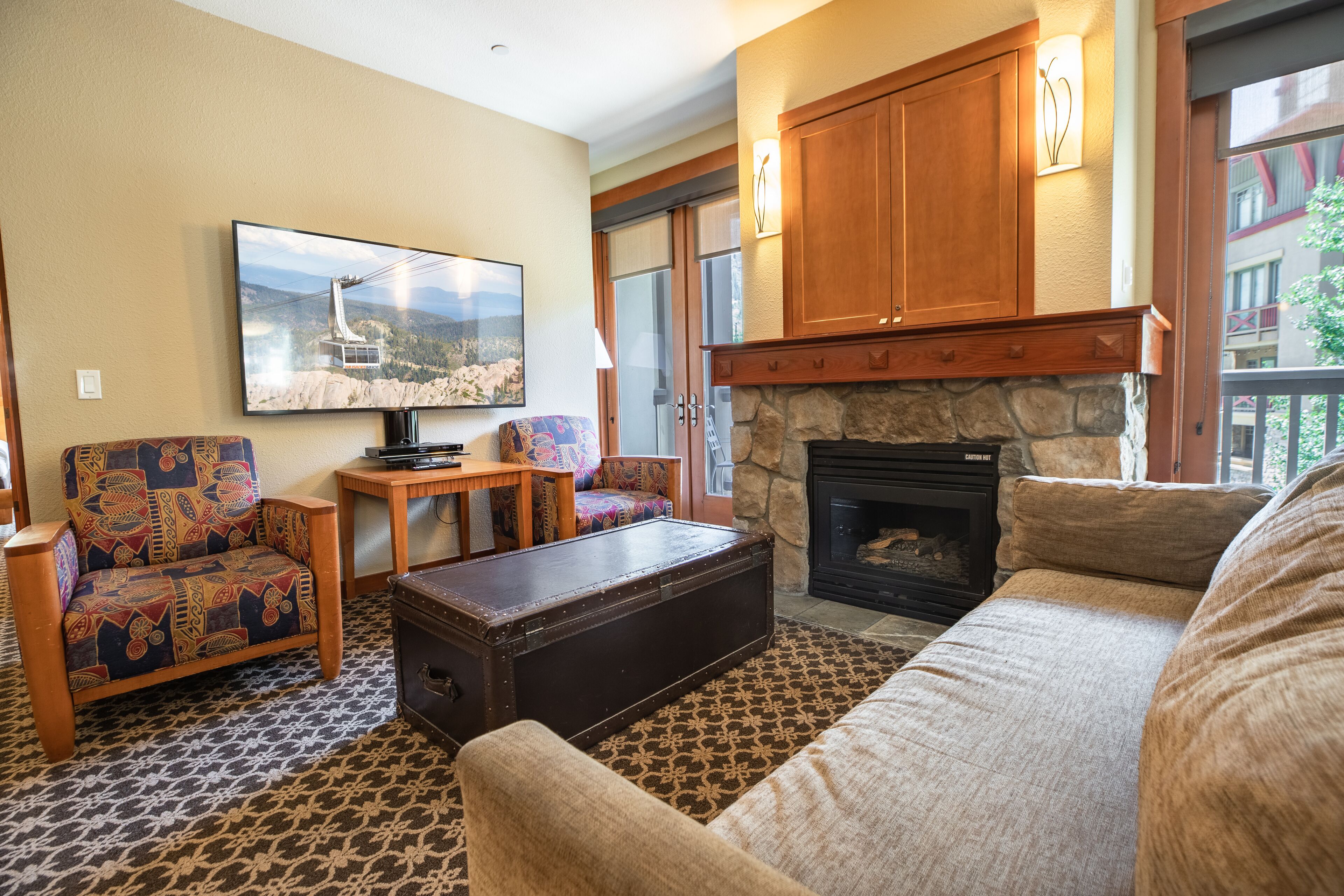 A cozy living room with a stone fireplace, a large flat-screen TV, patterned carpet, and a colorful armchair.
