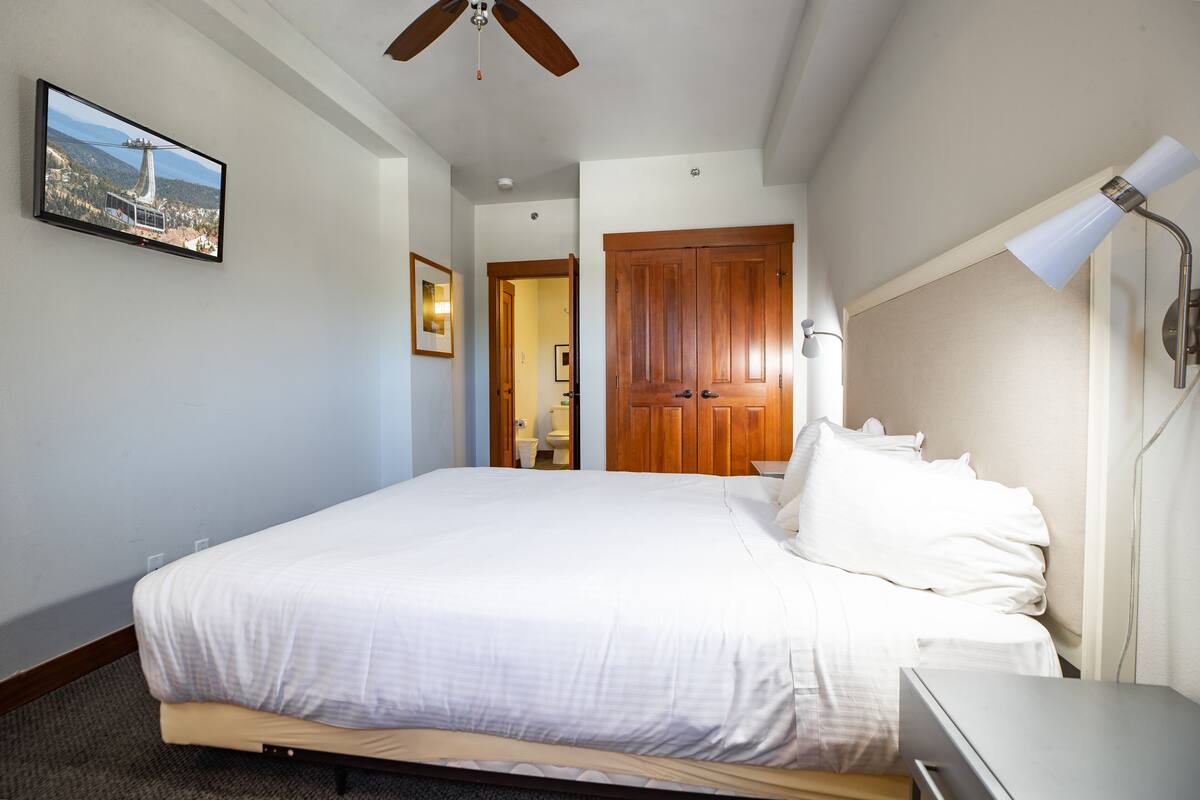 A hotel room with a large bed, a ceiling fan, a TV mounted on the wall, and a wooden door.
