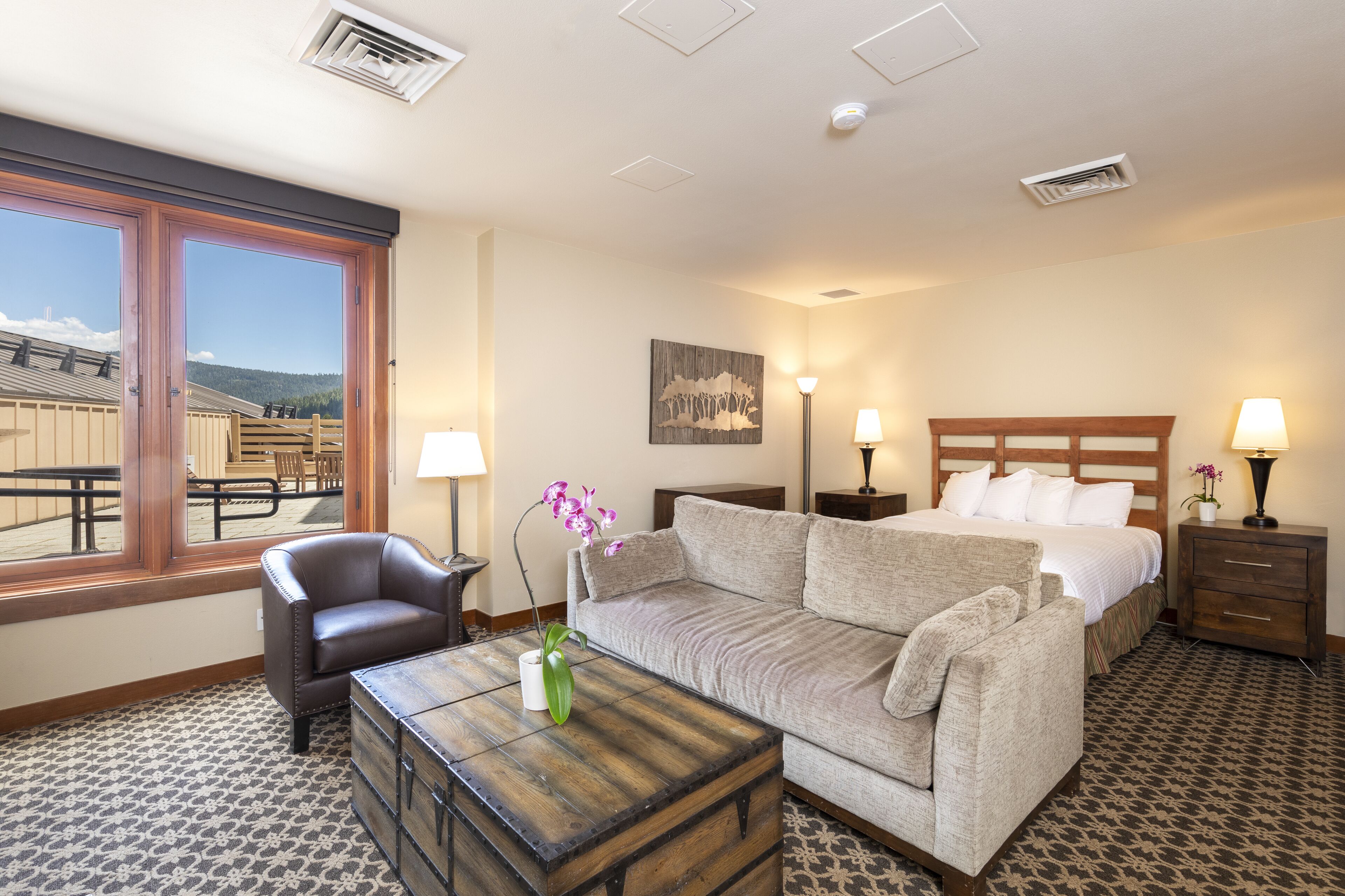 Habitación de hotel con cama, sofá, sillón, un cofre de madera y ventana con vistas a un edificio y árboles.