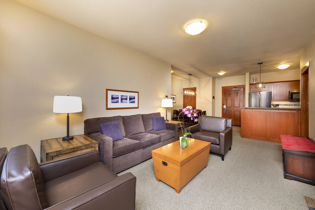 A hotel room with a living area featuring a sofa, a coffee table, and a lamp. There is a kitchen area with a refrigerator and a microwave.