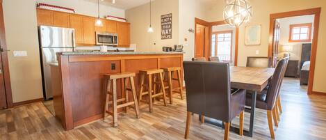 A modern kitchen with wooden cabinets, a central island, bar stools, and a dining area with a large table and chairs.