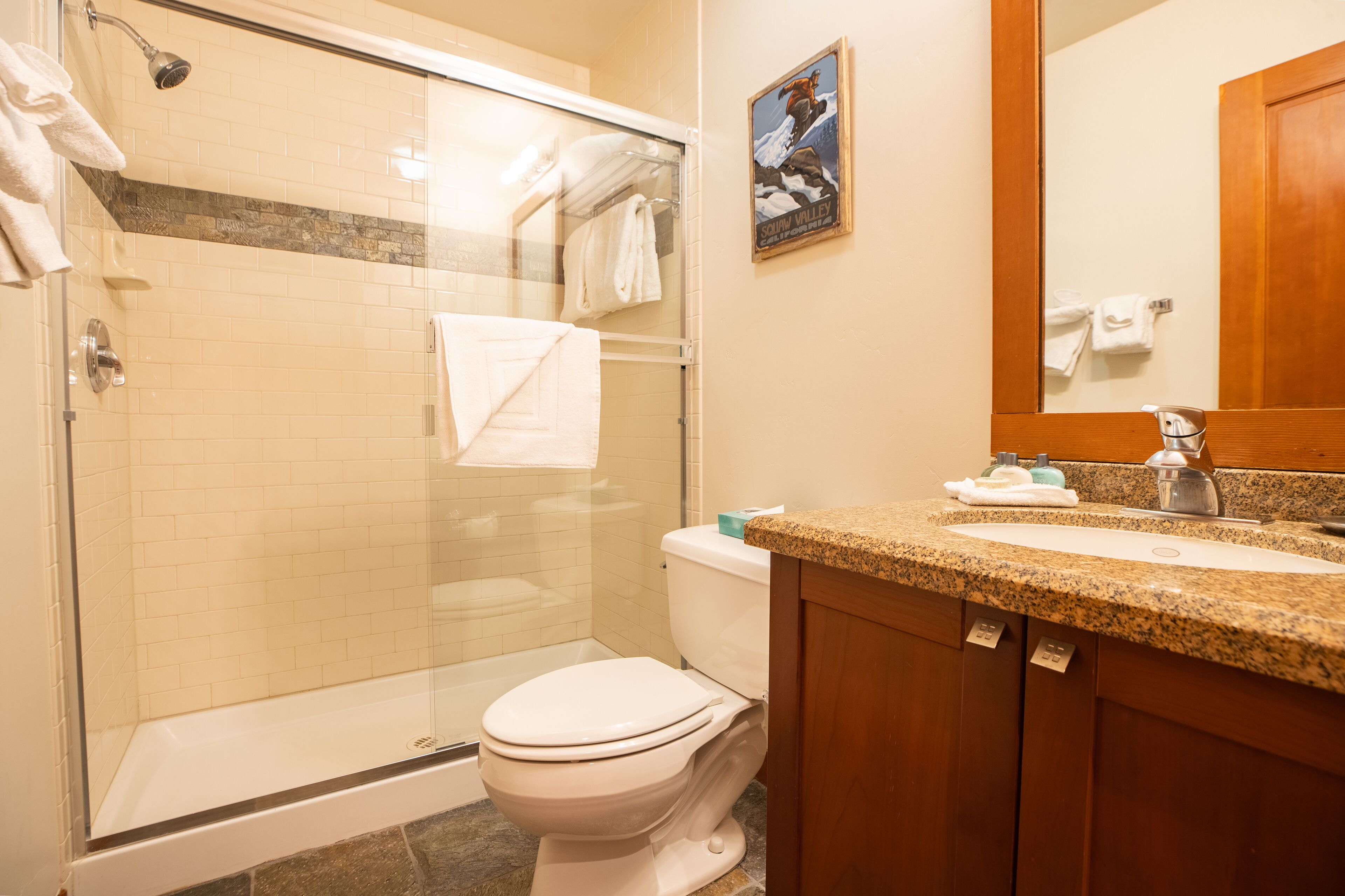 A bathroom with a glass-enclosed shower, a toilet, a sink with a granite countertop, and a wooden cabinet.